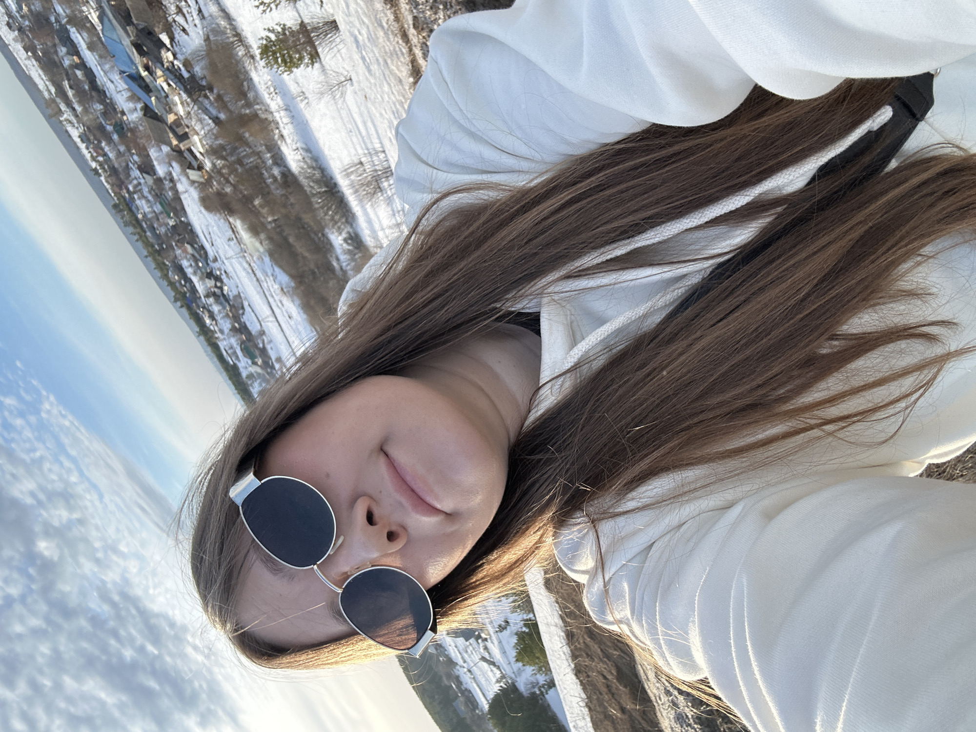 A female individual is depicted in a close-up, slightly tilted self-portrait, positioned against a wintery landscape identified as Фоки, Russia. The subject has long, brown hair and is wearing a white hooded sweatshirt with visible white drawstrings. Silver-framed sunglasses with dark lenses obscure her eyes. Her nose, mouth, and chin are visible, with her lips appearing closed. A black strap, possibly from a bag or device, is visible across her chest beneath her hair. She maintains direct eye contact with the camera. The background presents an expansive view of a snow-covered terrain, featuring a rural settlement with numerous houses of varying roof colors, sparse vegetation, and bare trees. The sky is partially cloudy with patches of blue. Strong, low-angle sunlight illuminates the scene, casting highlights on the subject and the snowy landscape.