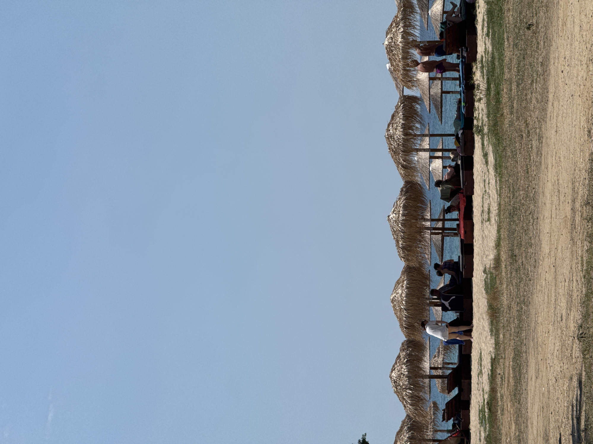 The image depicts a beach scene with a row of thatched umbrellas providing shade. Several people are visible relaxing under the umbrellas or standing nearby. The sea is a calm blue, and the sand is a light tan color, with some sparse green vegetation bordering the sandy area. The sky is clear and a light blue, suggesting a sunny day. The time of day appears to be daytime, with the sun casting shadows on the sand. The overall mood is one of relaxation and leisure, typical of a beach vacation. The people are dressed in casual beachwear, indicating a warm climate. The scene is tranquil, with no obvious activity other than people enjoying the beach. There is no visible text in the image.