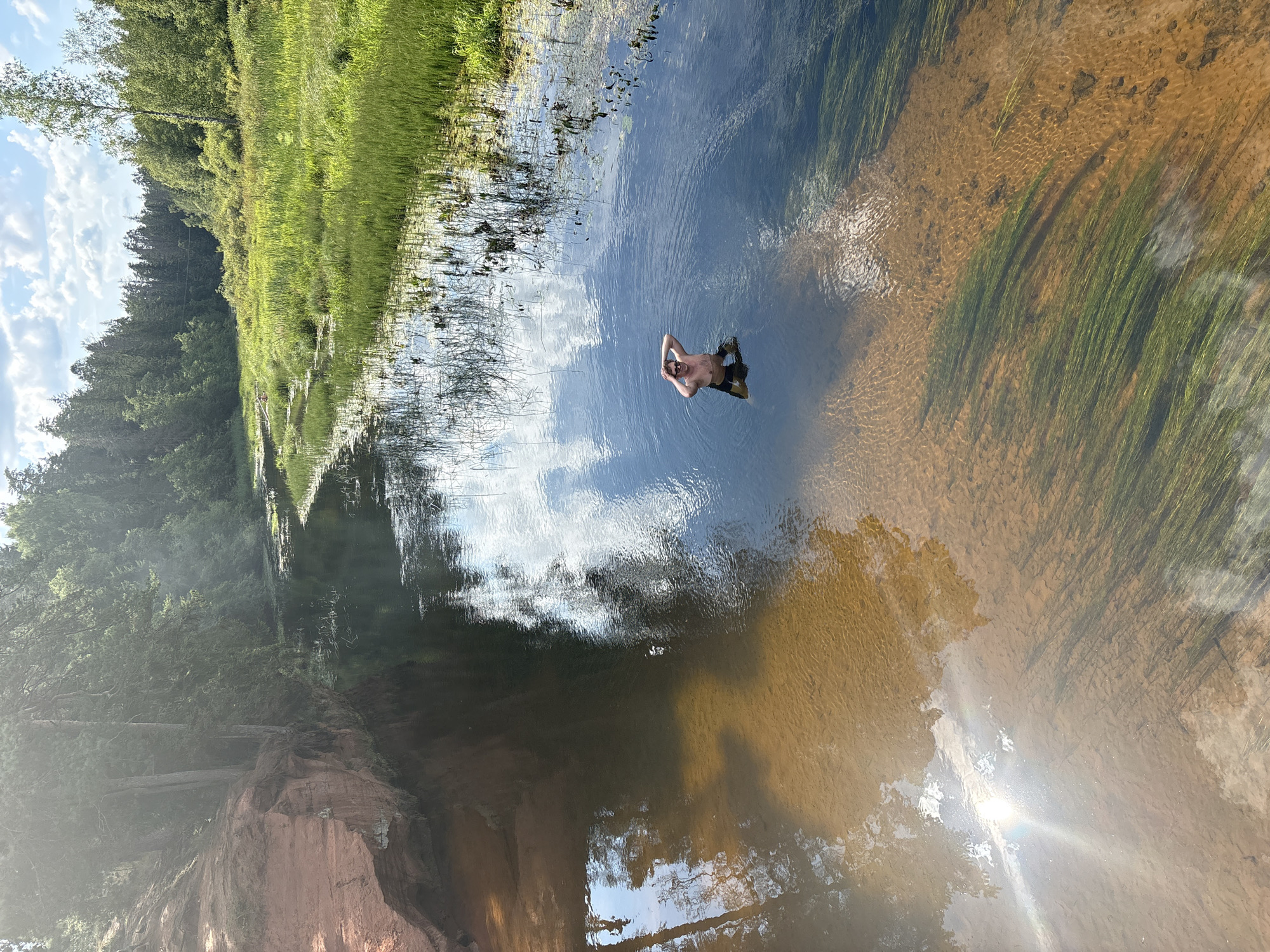 The image depicts a serene river scene in Dymishche, Russia. A man is standing in the shallow water of a clear river, with his hands clasped behind his head and wearing dark swim trunks. The riverbed is visible, appearing sandy with patches of submerged aquatic vegetation. To the left, a reddish-brown rocky cliff rises from the water's edge, covered in trees. The river flows into a wider expanse of water on the right, bordered by a dense green forest and lush, tall grass on the banks. The sky above is a mix of blue with scattered white clouds, reflecting on the water's surface. The overall atmosphere suggests a tranquil, natural setting on a bright, sunny day.