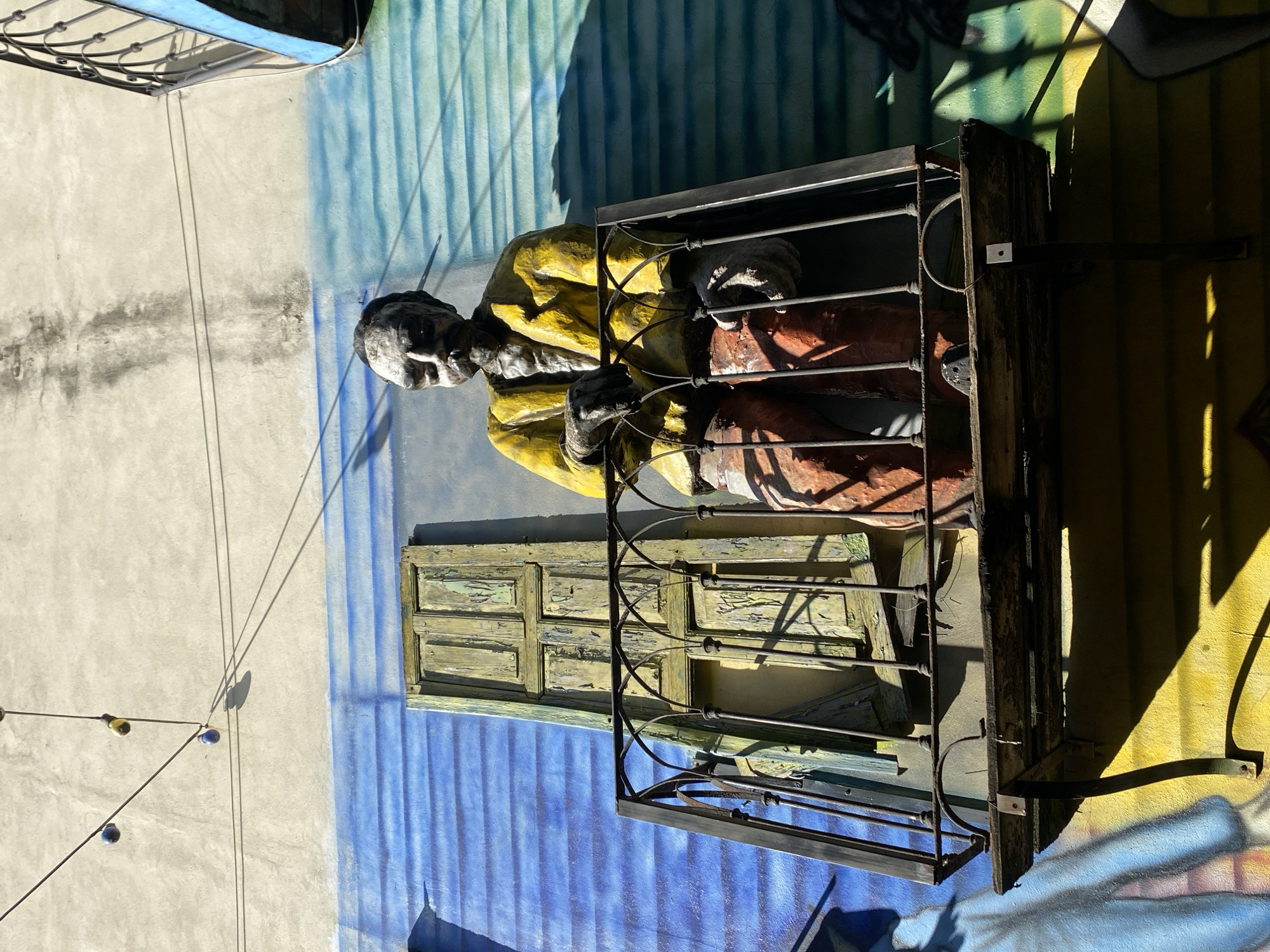 This image depicts a vibrant outdoor scene in Buenos Aires, Argentina, featuring a life-sized figure positioned against a colorful wall. The central subject is an older man, appearing as a statue or mannequin, with a white or grey head. He is dressed in a bright yellow top, possibly a poncho or jacket, and reddish-brown trousers, and is seated or leaning within a rustic, dark metal balcony-like structure made of wood and metal railings. His hands rest on the railings.
Behind the figure, an old, weathered green wooden window with peeling paint is visible. The background wall is a prominent feature, painted in distinct sections: a light grey-beige on the far left, transitioning into a bright blue section painted to resemble corrugated metal, and then a sunny yellow section on the right, which also features elements of colorful graffiti or a mural at its lower edge.
The scene is illuminated by bright, direct sunlight, casting long, sharp shadows from the figure, the balcony, and a string of lights. These string lights, adorned with small yellow and blue bulbs, are stretched across the grey-beige wall on the left. The distinct shadows indicate a clear, sunny daytime, likely around mid-day. The overall ambiance is artistic and characteristic of urban street art. There is no visible text in the image.