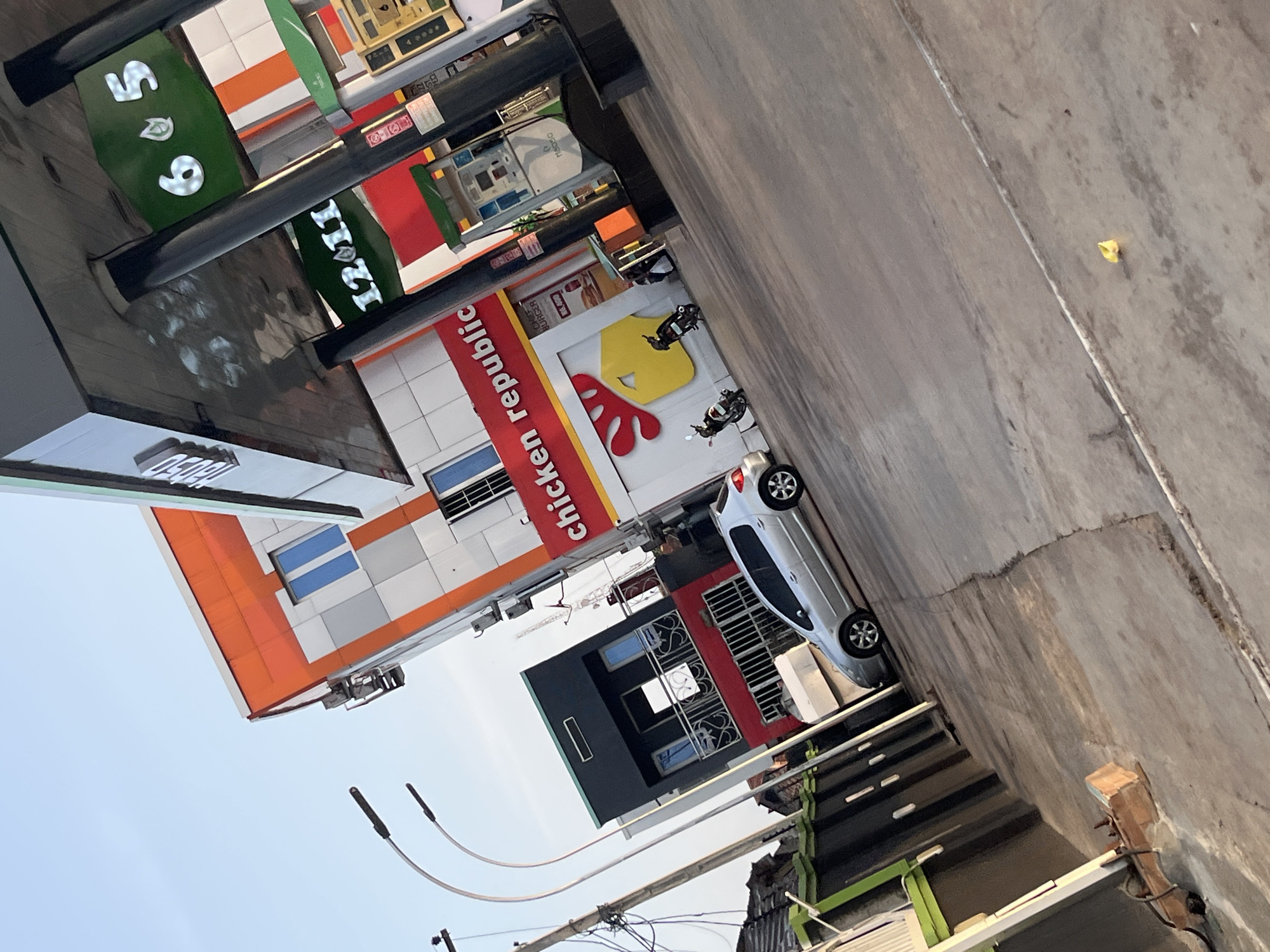 The image shows a view of a gas station and adjacent buildings in what appears to be a city. The gas station has several pumps under a large overhang supported by black pillars. Illuminated signs on the overhang display prices such as "65" and "12.01". A building with a prominent red sign that says "CHICKEN REPUBLIC" is visible behind the gas pumps, featuring a yellow chicken logo with a red comb. To the left of the gas station, there is a multi-story building with a distinctive orange and white facade, and a black and red building with balconies. A silver hatchback car is parked on the asphalt in front of the buildings. Two motorcycles are also visible, one parked near the Chicken Republic building and another one in motion. A street light with two lamps curves over the scene on the left. The ground is paved with concrete, showing some cracks and markings. The sky above is clear and blue.
