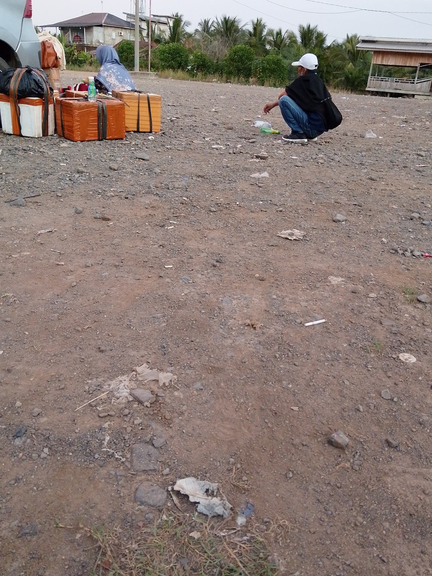 In Muara Sungsang, Indonesia, a man wearing a white cap and a dark shirt squats on a gravelly ground, facing away from the camera. He has a dark bag slung over his shoulder. In front of him, several pieces of luggage are arranged. To the left of the man and closer to the foreground, there is a stack of wrapped packages, some appearing to be cardboard boxes covered in orange plastic and secured with black straps. One package is white with black straps, and another appears to be a black duffel bag. To the right of these packages, a person wearing a blue headscarf is seated. In the background, there is a building with a brown roof, some palm trees, and a wooden structure that appears to be on stilts. The ground is rough and uneven, with scattered small stones and some debris. The sky is overcast.