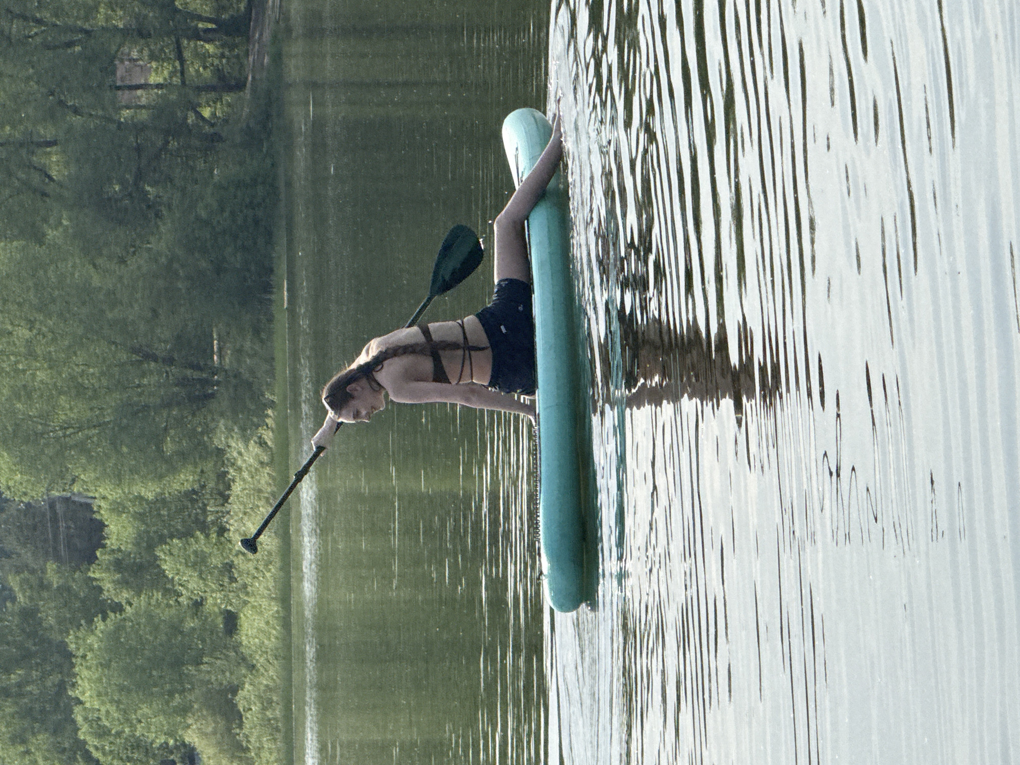 A woman is on a light blue paddle board in the water. She is leaning over, placing a black paddle into the water. The surrounding water is slightly disturbed with reflections. Trees are visible on the bank to the left. The scene appears to be Novosinkovo, Russia.