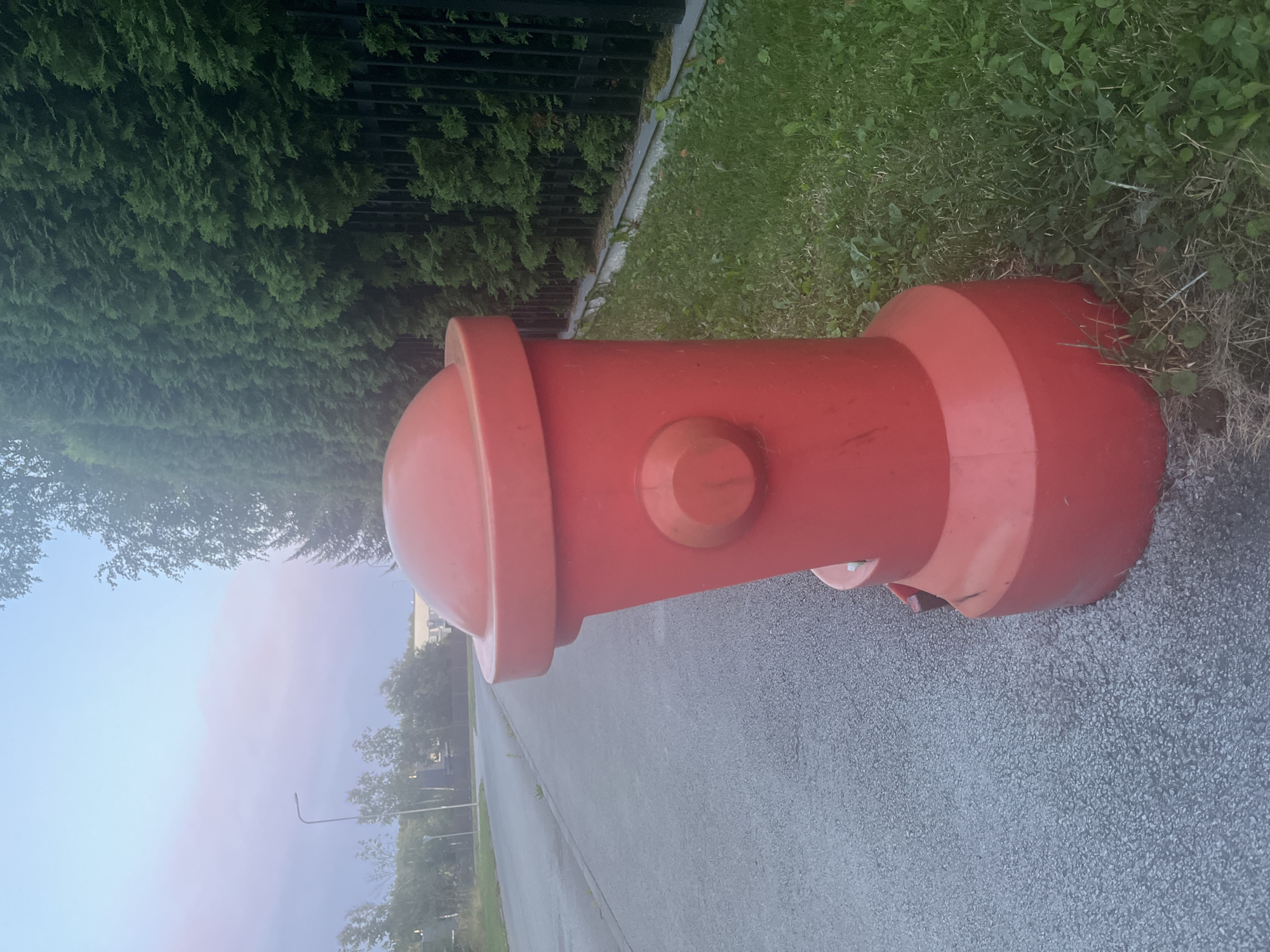 The image displays a bright red, plastic fire hydrant placed on the side of an asphalt road. The hydrant is positioned in the foreground, taking up a significant portion of the right side of the frame. It is a standalone object with a rounded top and a cylindrical body featuring a circular protrusion on its side. The road extends to the left and into the background, appearing to be a street in an urban or suburban setting.
To the right of the hydrant, there is a strip of green grass, and behind the grass, a dense, tall hedge of dark green evergreen trees is visible. A black metal fence runs along the base of the hedge. In the background, beyond the road, there are trees and some buildings, suggesting a developed area. The sky above is a pale blue with hints of pink and purple, indicating either early morning or late evening light.
The scene is devoid of any people or animals. The overall impression is one of a quiet, possibly residential or commercial street, with the fire hydrant serving as a prominent, artificial element in the natural and man-made environment. The location is identified as Haabneeme alevik, Estonia.