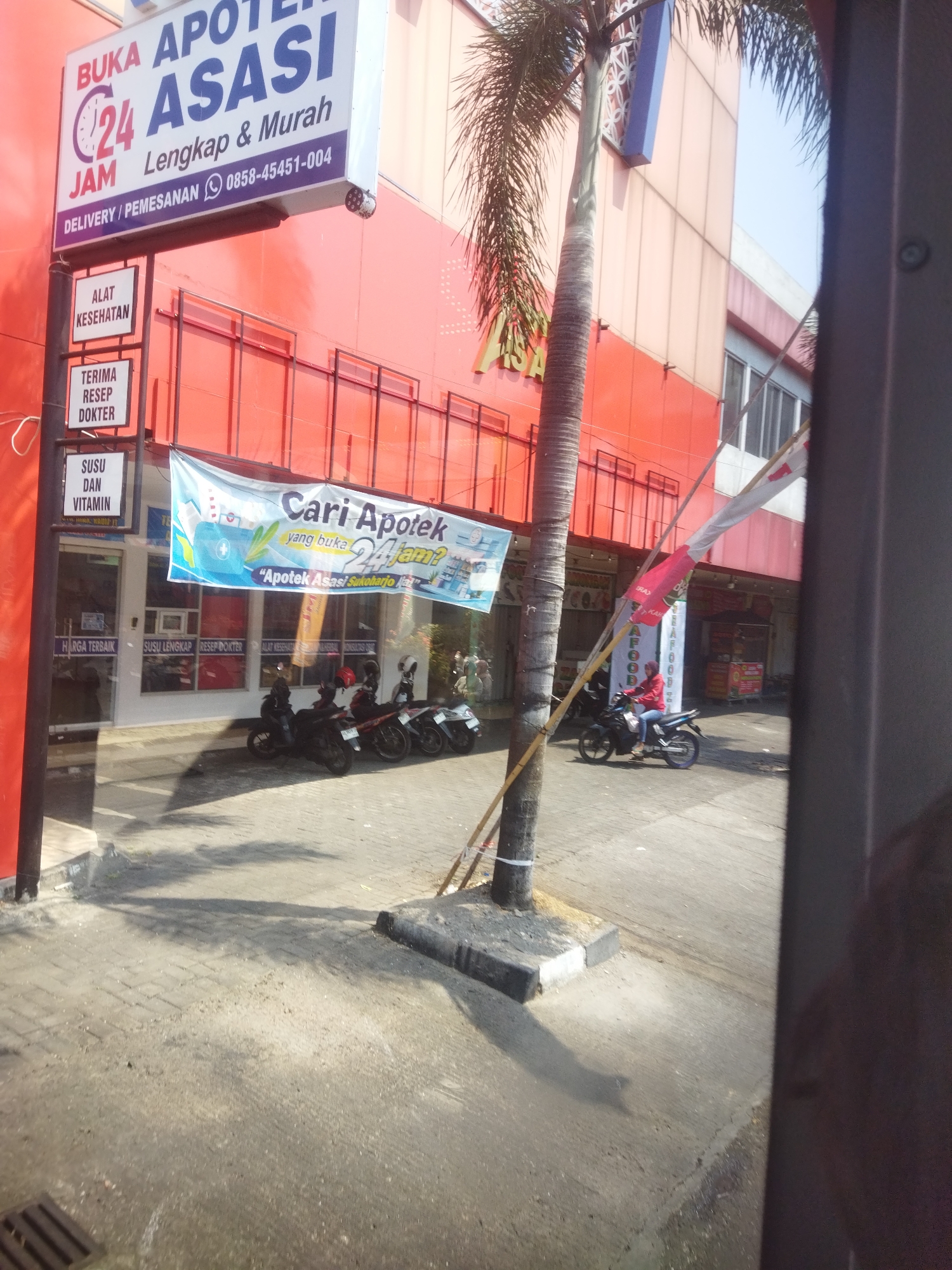 The image shows the exterior of an "Asasi Apotek" (Pharmacy) in Sukoharjo, Indonesia. The pharmacy is part of a building with an orange facade. A prominent sign above the entrance reads "BUKA APOTEK ASASI 24 JAM Lengkap & Murah" (Open Pharmacy Asasi 24 Hours Complete & Cheap). Below this, there are smaller signs indicating "ALAT KESEHATAN" (Health Equipment), "TERIMA RESEP DOKTER" (Accepts Doctor's Prescriptions), and "SUSU DAN VITAMIN" (Milk and Vitamins). A banner in front of the pharmacy reads "Cari Apotek yang buka 24 Jam?" (Looking for a pharmacy open 24 Hours?).
Several motorcycles are parked in front of the pharmacy. In the background, a person on a motorcycle is seen riding away. A tall palm tree stands to the left of the pharmacy entrance. The ground is paved with interlocking bricks. The scene appears to be during daylight hours, with shadows cast on the pavement.