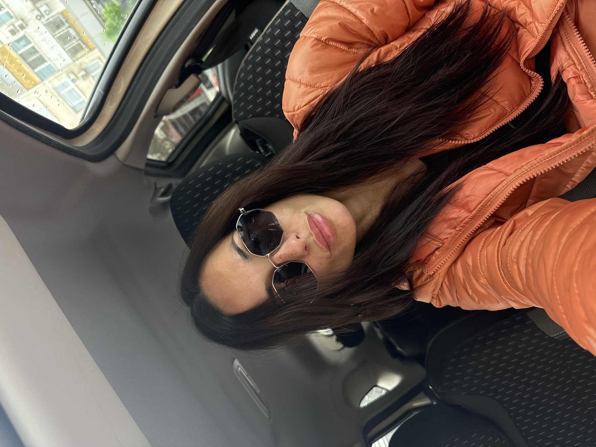 A woman with long, dark brown hair, wearing sunglasses and an orange puffer jacket, is positioned in the driver's seat of a car. The car is located in Pleven, Bulgaria, based on visible buildings outside the window. The image is angled, presenting a skewed view of the car's interior. Raindrops are visible on the car window.