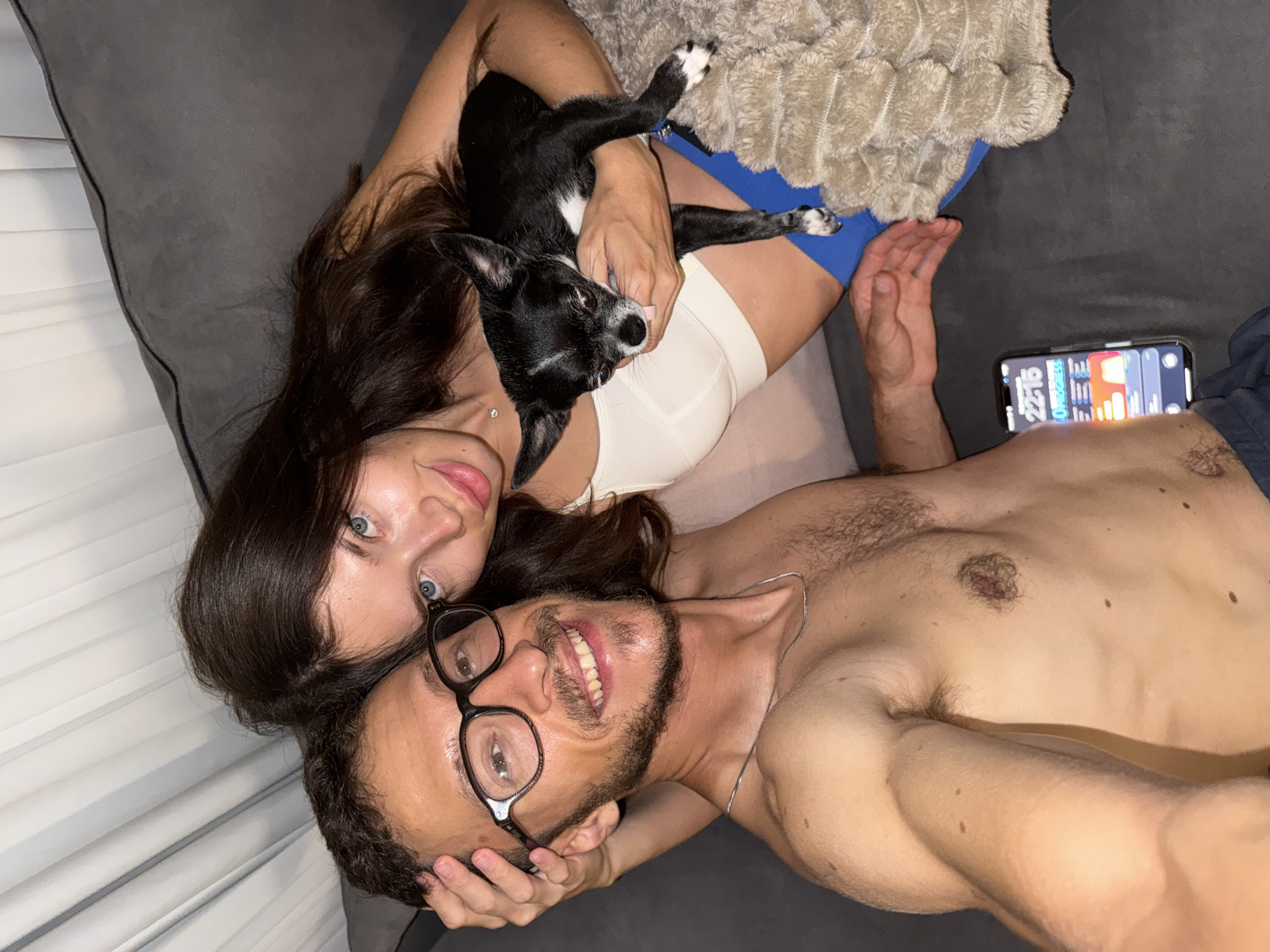 A man and a woman are pictured embracing on a couch with a small black and white dog. The man, wearing glasses, has his arm around the woman, and she is holding the dog. A mobile phone with a colorful display is visible on the couch. The scene takes place indoors, likely in a residential setting. There is no visible indication of the city of Um Rummane, Jordan.