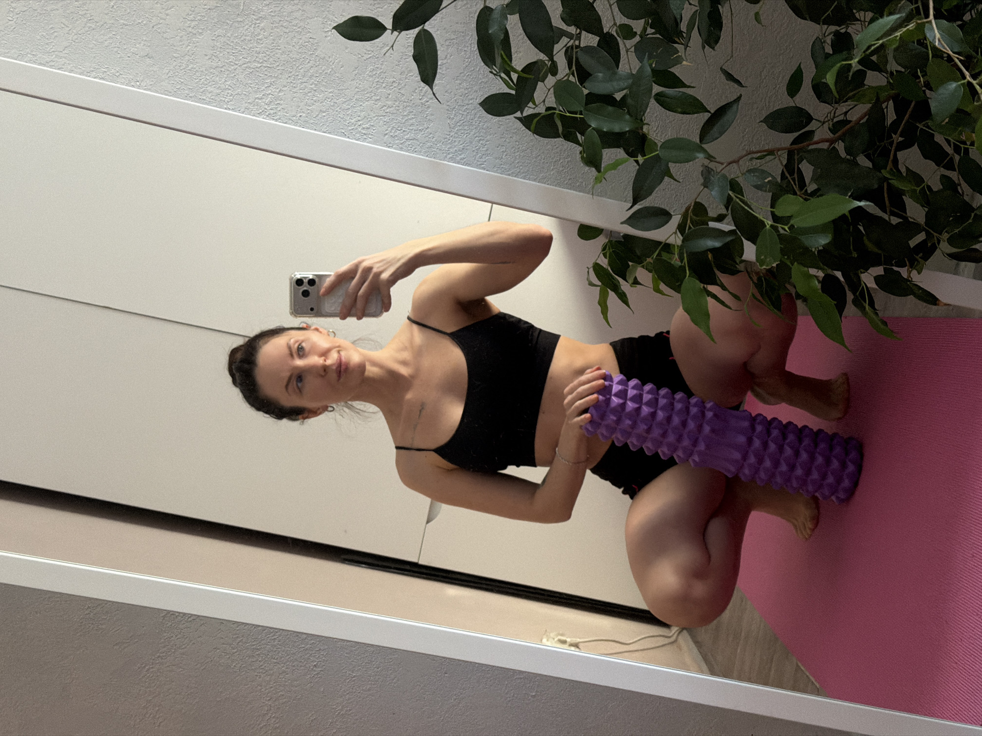 A woman is crouching on a pink yoga mat in what appears to be an apartment in Perm, Russia. She is holding a purple foam roller. She is wearing a black athletic crop top and black shorts. She is taking a selfie with her smartphone in front of a mirror. Behind her is a white wardrobe and a potted plant with green leaves.