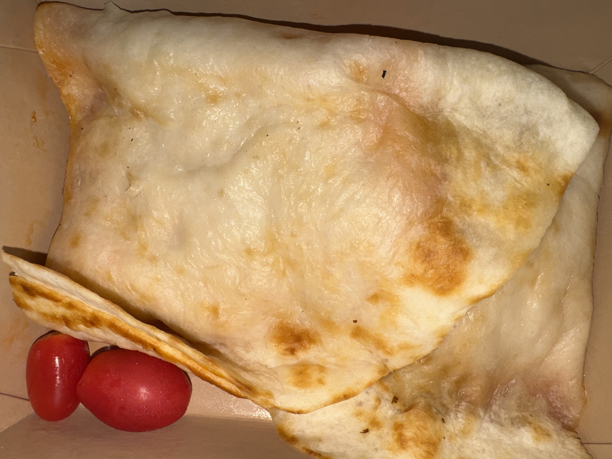 Two ripe cherry tomatoes, one oval and one spherical, rest at the upper left of the frame. To their right, a folded piece of flatbread, golden brown with some charred spots, is the primary subject. The flatbread appears to be part of a larger dish, as another folded piece is visible beneath the main portion. The food is contained within a light brown cardboard box. The scene is an overhead shot of food in a container.