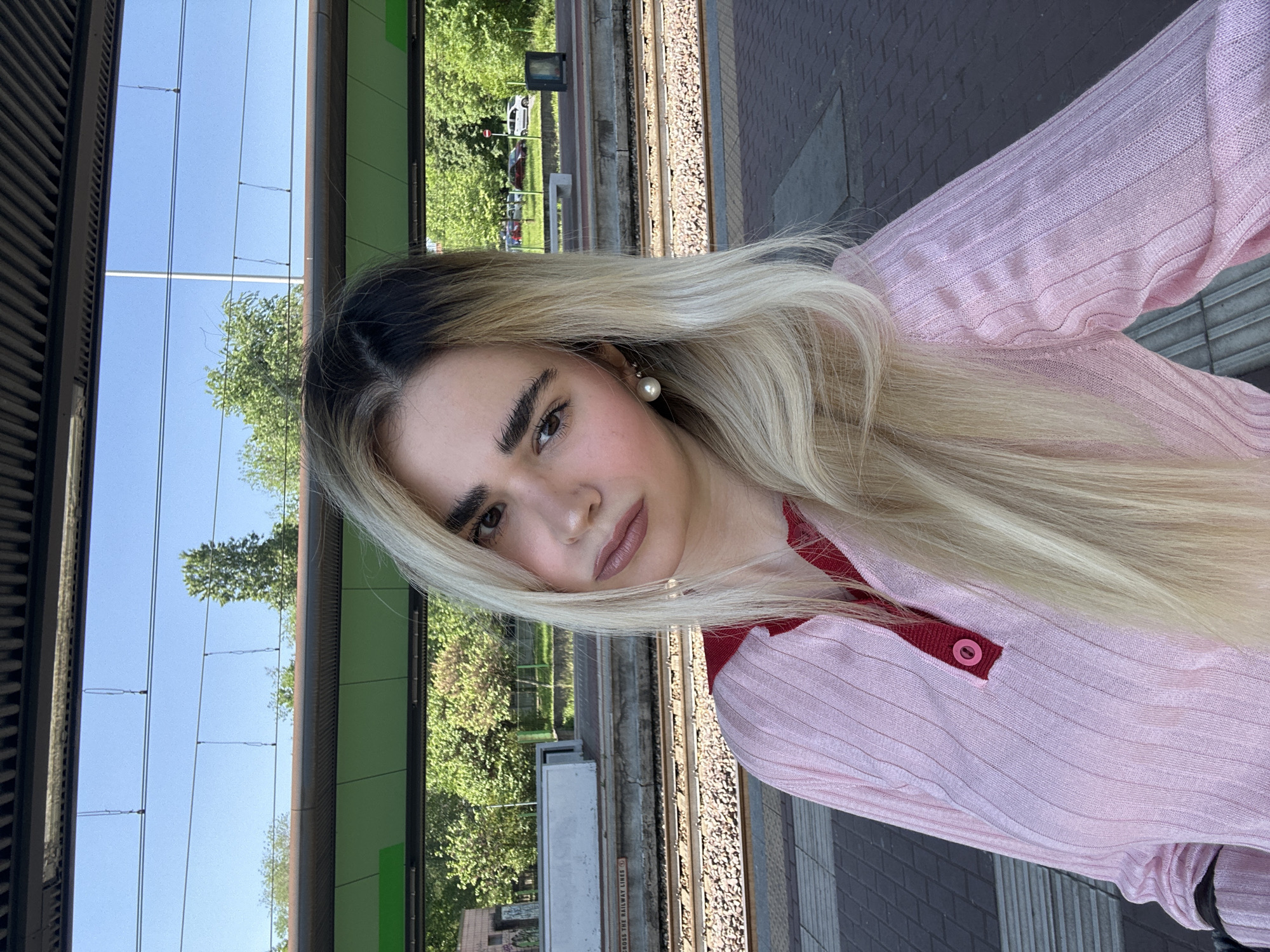 A woman is standing on a train platform. She has fair skin, dark eyebrows, and long blonde hair. She wears a pink sweater with red accents. The platform is made of gray pavers. The tracks run alongside the platform. In the background, there are trees, buildings, and a blue sky. The photo was taken outdoors during the day. Based on the architecture and train platform design, the location could be in Milan, Italy.