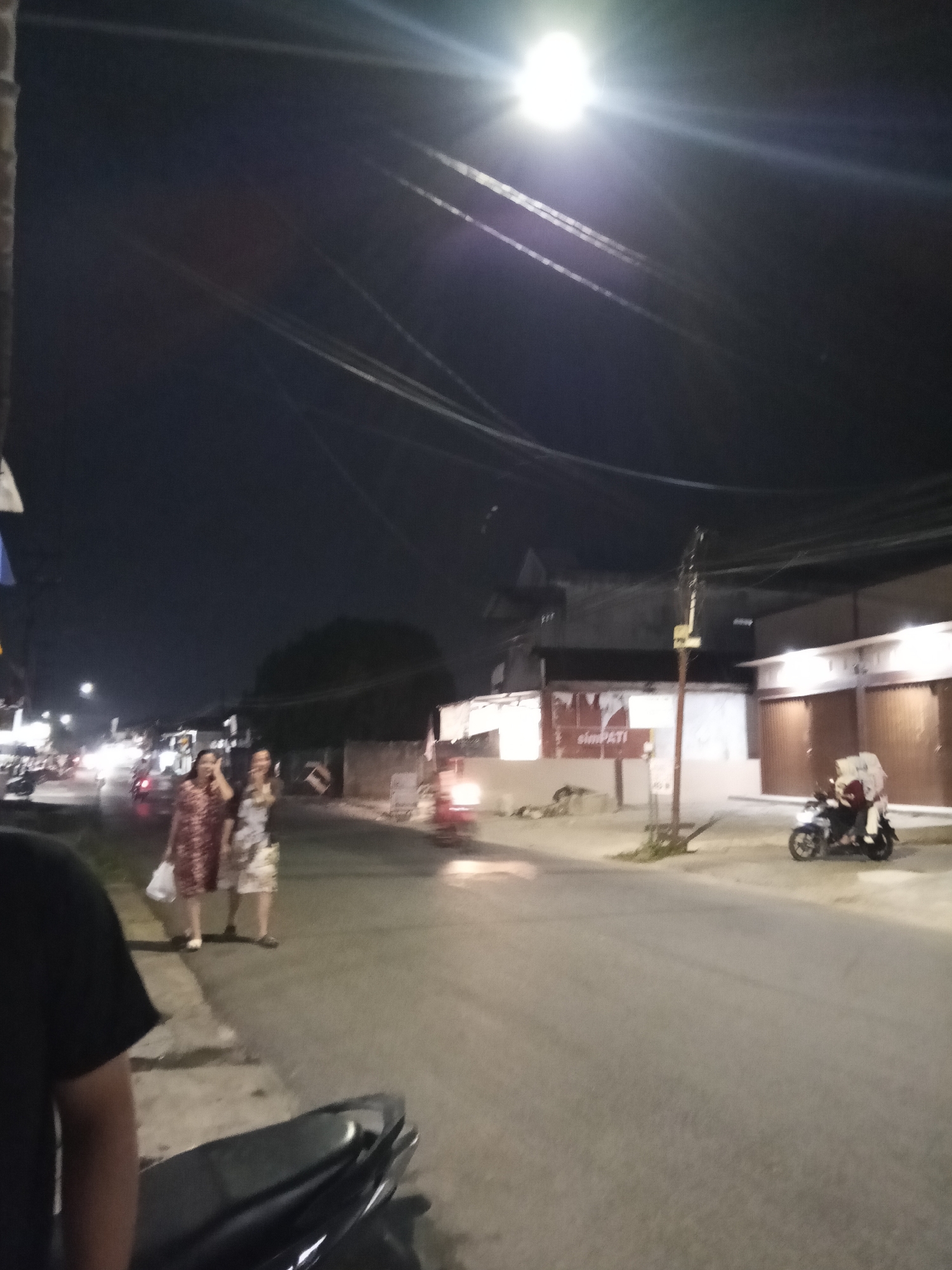 At night, on a paved street in what appears to be a commercial area in Lubuklinggau, Indonesia, two women walk on the sidewalk. The woman on the left wears a red and white patterned dress and carries a white plastic bag. The woman on the right wears a lighter-colored, patterned dress. They are walking away from the camera and appear to be interacting. Further down the street, a motorcycle with two riders, one in a white hijab, is visible on the road. Several buildings line the street, some with illuminated signs and closed shutters. Overhead, power lines are strung across the dark sky, illuminated by a bright street lamp in the upper left quadrant of the image. The ground is asphalt, and there are indications of a concrete curb and sidewalk. The overall scene depicts a common street view at night, likely in a moderately populated urban area.