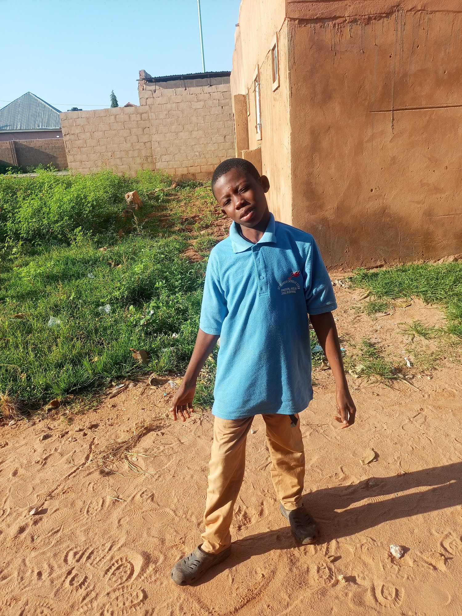A young Black boy stands outdoors in a sandy area. He is wearing a light blue polo shirt with some writing on it and khaki pants. He is looking at the camera with a neutral expression. Behind him, there is a wall of a brown building. The ground is sandy with some grass growing to the left. The sky is clear and blue. The boy's shadow is cast to his right on the sandy ground. The image appears to have been taken in Tempure, Nigeria.