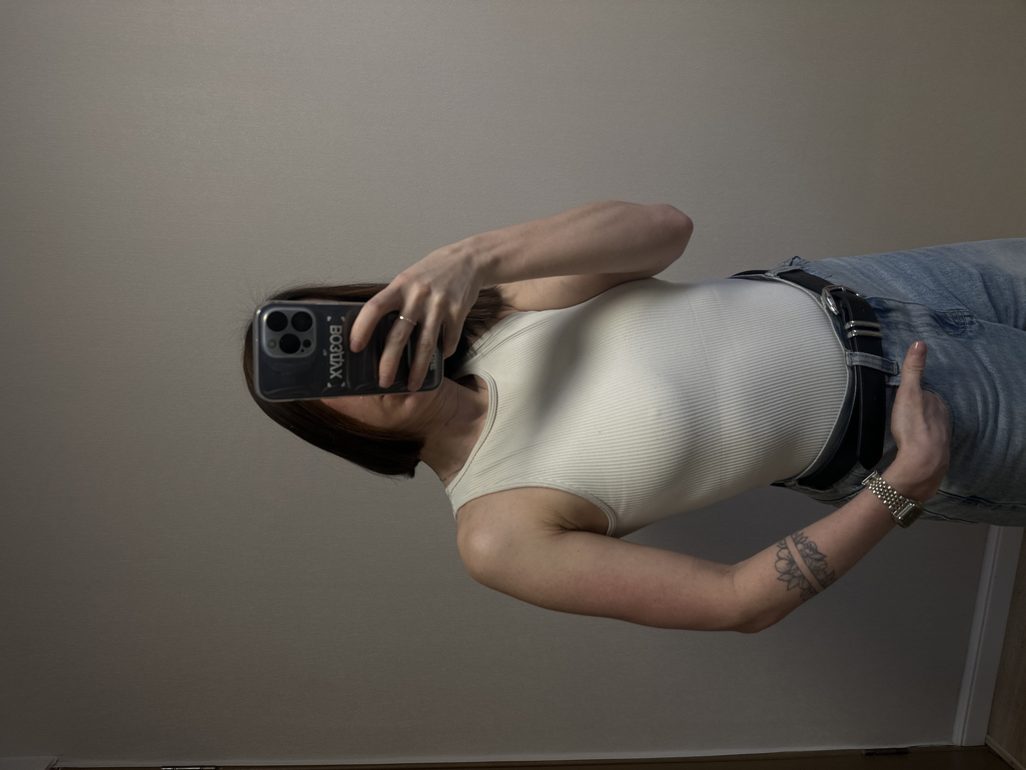 A woman is taking a selfie in front of a light gray wall. She is wearing a white ribbed tank top and light wash jeans with a black belt. She has a watch on her left wrist and a tattoo on her left forearm. The woman is holding a black phone with the brand name "BOZDAX" written in Cyrillic characters on the case.
There is no indication that this image was taken in Votkinsk, Russia.