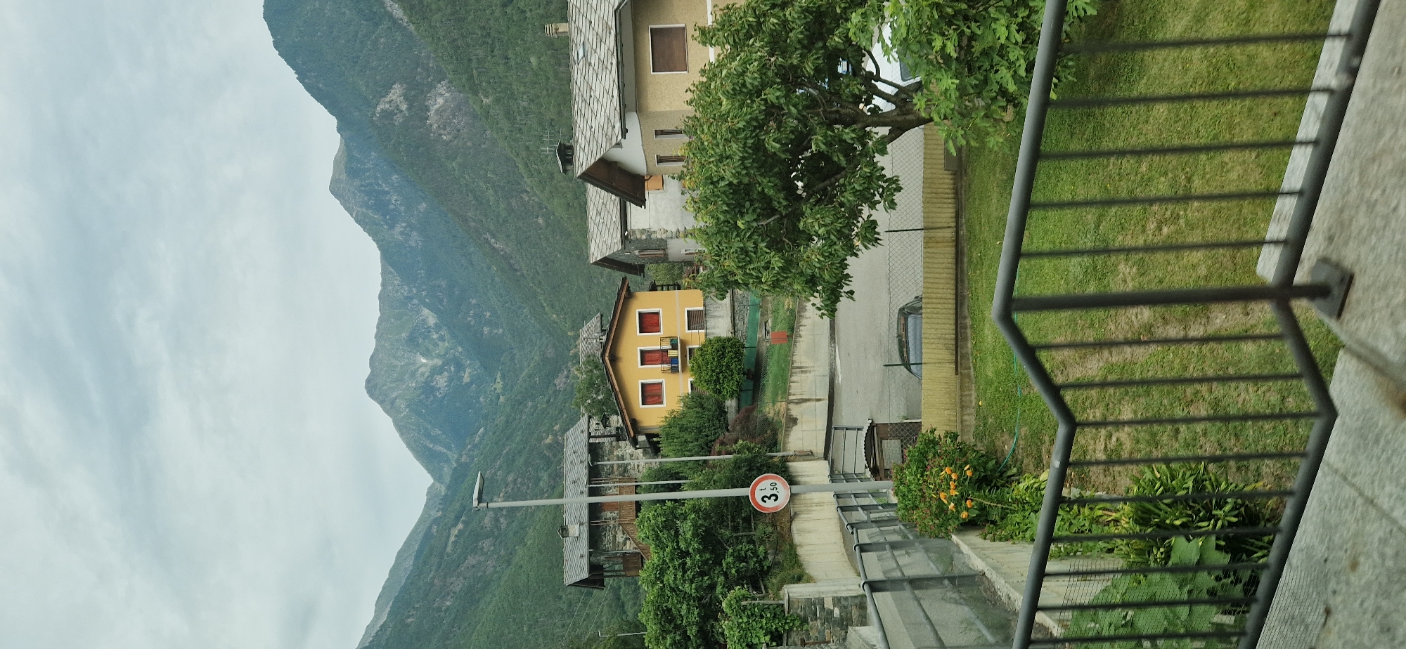 This is a landscape photograph taken outdoors, likely in a mountainous region given the prominent mountains in the background. The scene depicts a small village with several houses nestled on a hillside. The houses appear to be traditional in style, with pitched roofs and balconies. There's a yellow house with red window frames and balconies, and other houses with grey tiled roofs. Lush green trees and vegetation surround the buildings, suggesting a rural or semi-rural setting.
A road or pathway is visible, with a parked car. A speed limit sign with "3,50 t" is also present, indicating a weight restriction. The sky is overcast, suggesting a cloudy day. The overall atmosphere is peaceful and serene. The orientation of the image is portrait, with the top of the frame showing the sky and mountains, and the bottom showing the foreground with a railing and some foliage. The lighting suggests it is daytime, but not necessarily bright sunlight. The location is identified as Challand-Saint-Victor, Italy. There are no people visible in the frame, and the focus is on the natural scenery and the architecture of the village.