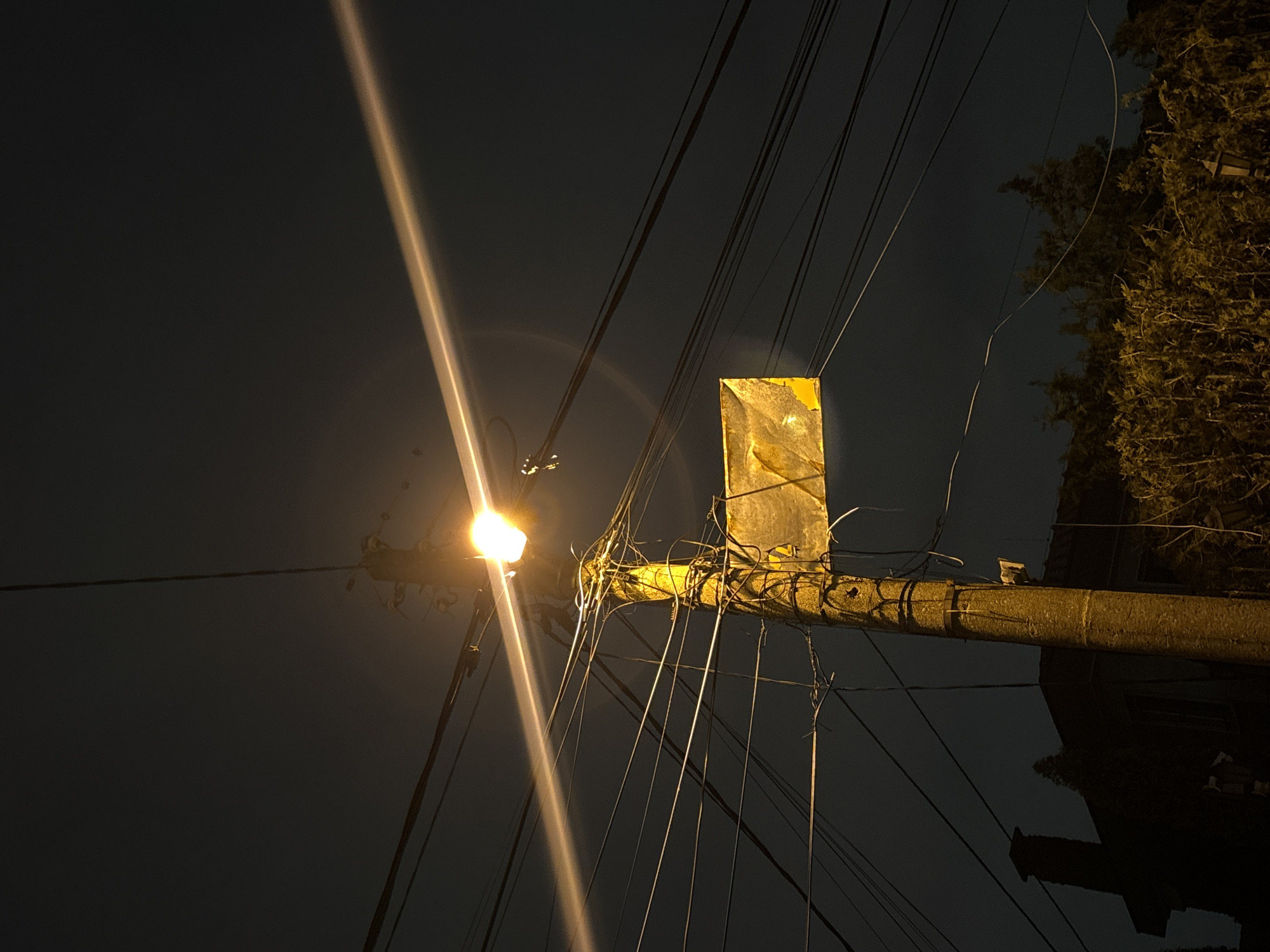 Captured at night, the image presents a low-angle upward view of a utility pole in an unidentified location in Sofia, Bulgaria. An incandescent light fixture emitting a bright, concentrated beam of light is mounted on the pole. The pole is draped with a dense web of electric wires and cables. A rectangular sign is attached to the pole above the light fixture. Foliage from trees is visible on the right side of the image. The sky is dark. The lighting conditions suggest that the utility pole is the primary source of illumination in the scene.