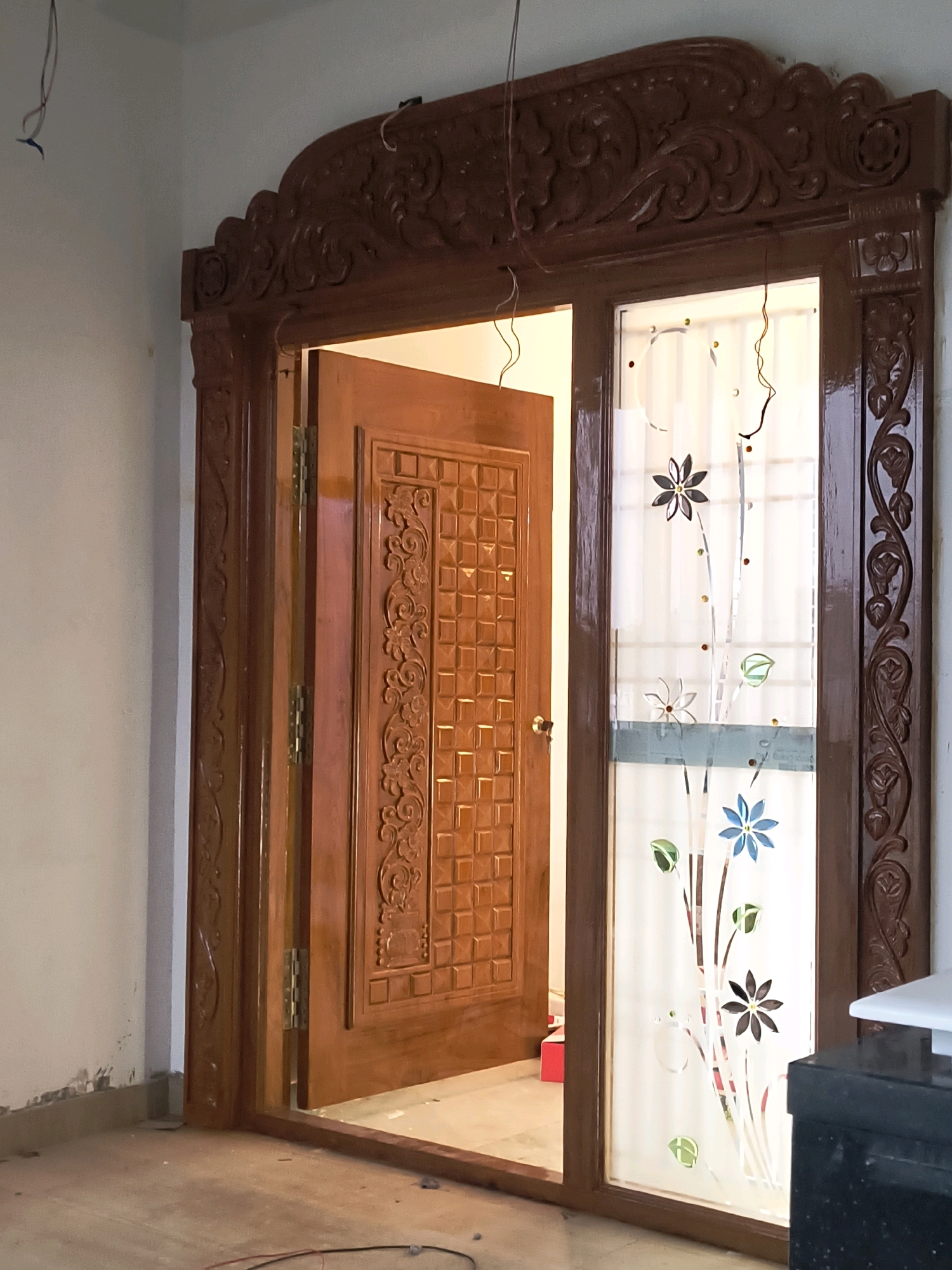 The image depicts an interior view of a wooden door frame and an adjacent glass panel with floral designs. The door frame is intricately carved with ornate patterns, featuring a decorative archway at the top. A wooden door with a patterned surface is ajar, revealing hinges. To the right, a glass panel is adorned with etched or applied floral motifs in shades of blue, brown, and green, with small colorful dots interspersed. The surrounding environment appears to be an indoor space under construction or renovation, with exposed electrical wires hanging from the ceiling and debris on the floor. The lighting suggests an indoor setting with natural light coming from the glass panel. The specific location in Ulvapadu, India, is not visually indicated in the image.