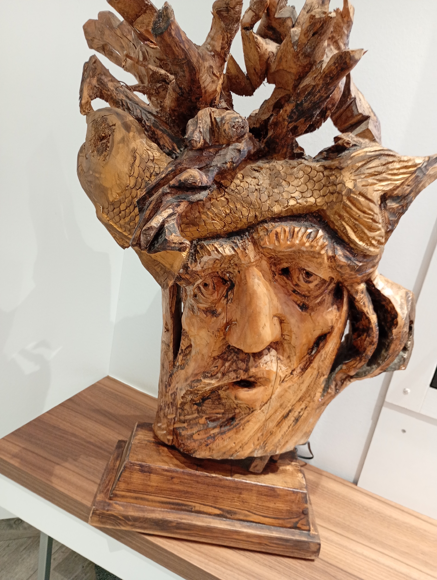 This image depicts a carved wooden sculpture of a face, possibly a bust, with intricate detailing and a rustic finish. The sculpture is mounted on a wooden base and rests on a wooden shelf against a white wall. The face appears aged, with deep-set eyes, a prominent nose, and a beard carved into the wood. A fish-like motif or texture is incorporated into the headpiece or headdress of the sculpture, with scales visible. The overall aesthetic suggests a handcrafted piece, possibly representing a mythical or historical figure. The lighting appears to be from artificial sources, casting subtle shadows. There is no discernible location within the city of Bad Sachsa, Germany indicated in the image.