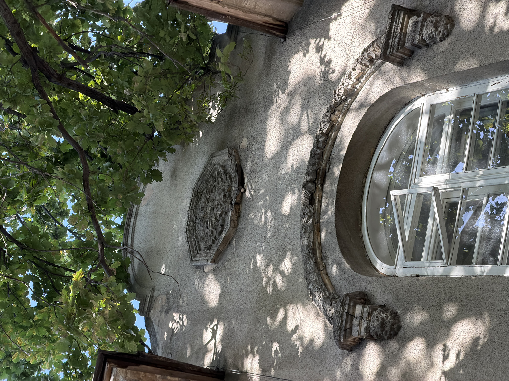A low-angle shot depicts the facade of a weathered building in Bishkek City, Kyrgyzstan, with a portion of a tree encroaching from the left. The facade is textured with a rough stucco finish, primarily gray in tone, and marked by shadows cast by foliage. On the wall, there is an octagonal, ornamental stone plaque, exhibiting intricate, perhaps floral or geometric, carvings. A decorative stone archway frames a rounded window with white mullions, which is partially open, revealing glimpses of the surrounding environment through the glass panes. The window frame appears aged and somewhat deteriorated. The architecture displays a blend of ornamentation and aging, indicative of a building with historical elements.