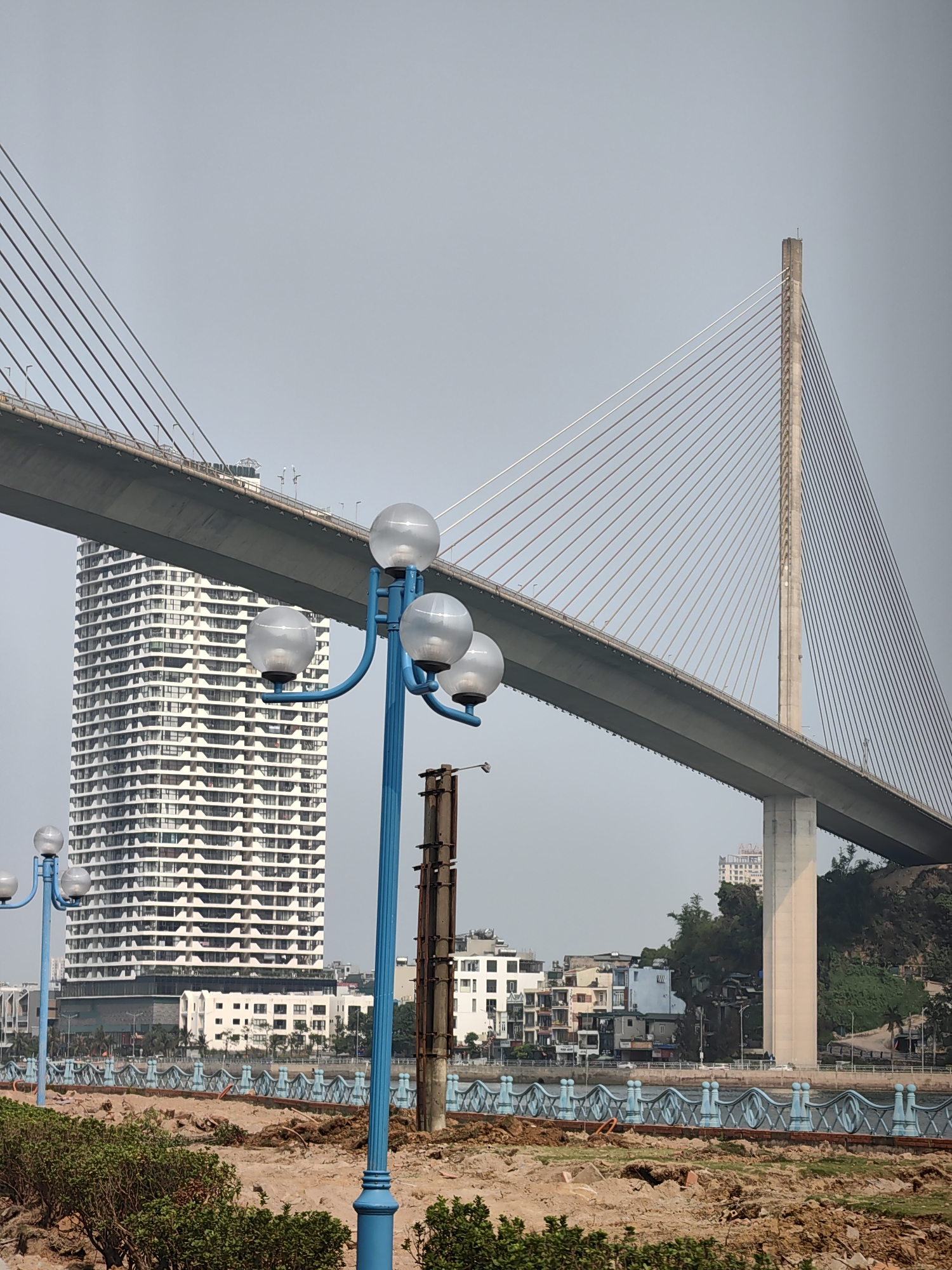 This is an exterior daytime shot of a large suspension bridge, probably the Bai Chay Bridge, as it appears to be located in Bai Chay Ward, Vietnam. A tall, modern, white building is visible to the left, partially obscured by the bridge. In the foreground is a recently disturbed patch of dirt and some plants, as well as a row of blue decorative fence sections and blue lamp posts. The sky is overcast.
