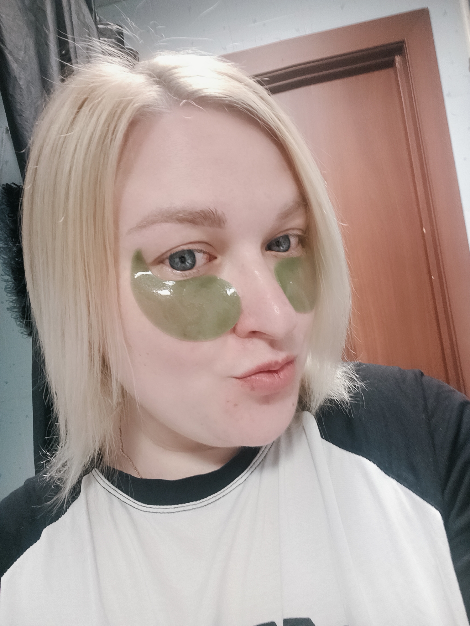 The media file presents a close-up, chest-up selfie of a young woman with platinum blonde, shoulder-length hair, situated indoors in Chaikovsky, Russia. She has fair skin and blue-grey eyes, which are directed at the camera. Applied beneath each eye are translucent, shiny green hydrogel patches, indicating a skincare routine or relaxation. Her lips are slightly pursed in a playful or pouting expression.
The woman is wearing a casual raglan-style t-shirt, characterized by a white body and black sleeves, with black stitching visible around the collar. A thin, gold-colored chain necklace is faintly visible around her neck. Her blonde hair shows some flyaways, particularly near the roots and along the ends.
The background elements suggest an indoor setting, possibly a bathroom or bedroom. To the left, a dark, textured curtain or fabric is partially visible. On the right, a segment of a light brown or wooden door frame is present. The lighting is soft and even, typical of an indoor environment, making the specific time of day unidentifiable. No visible text is present in the image. The overall mood conveyed is casual and relaxed.