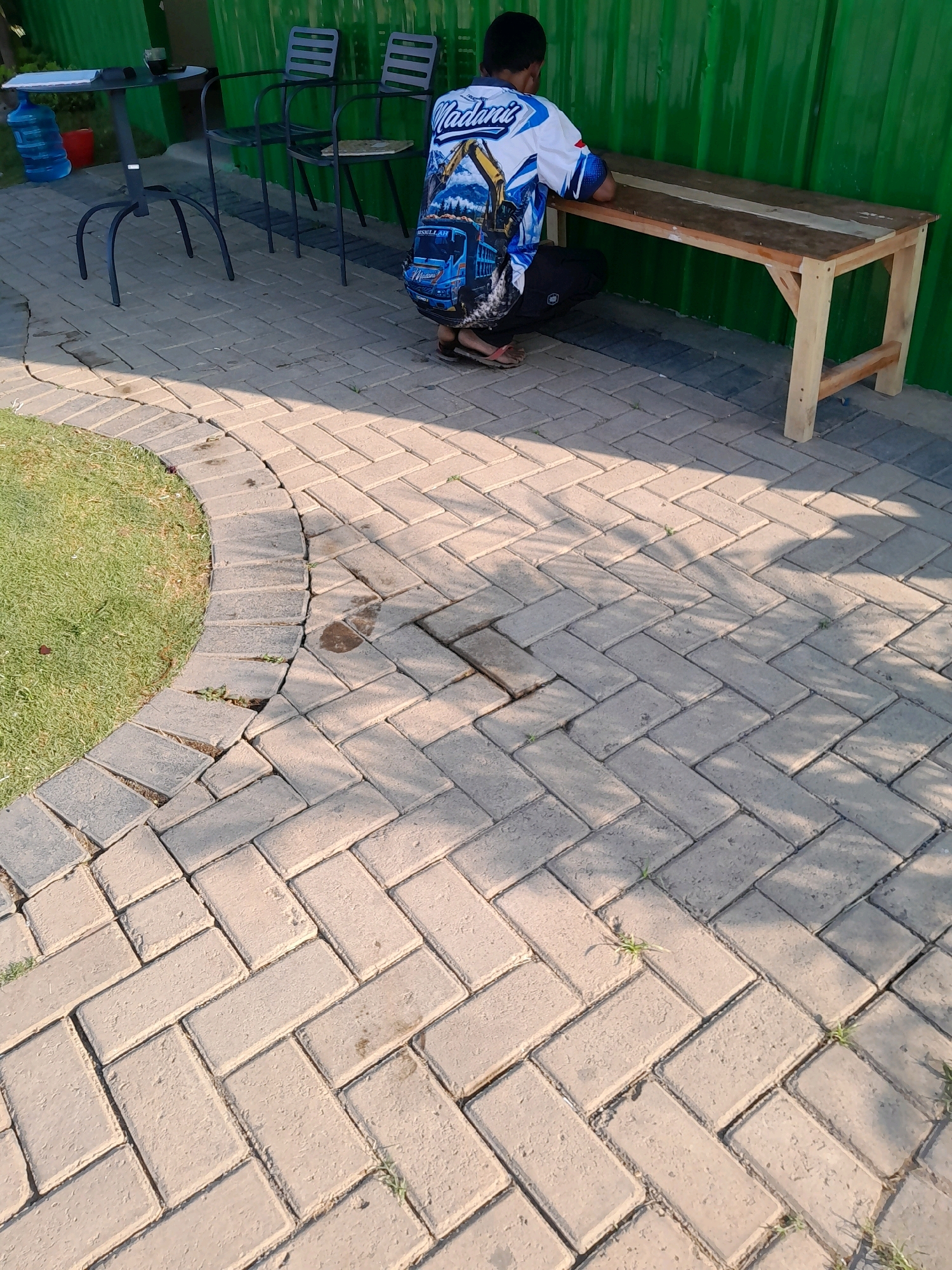 In Pakisaji, Indonesia, a person is seen crouching beside a wooden bench in an outdoor area. The person is wearing a white and blue shirt with a graphic design and black pants. They are facing away from the camera, towards the bench. The ground is paved with interlocking rectangular bricks arranged in a herringbone pattern. To the left of the paved area, there is a patch of green grass. In the background, there are two metal chairs and a tall, thin table with water bottles and a red bucket. A green corrugated metal fence forms the backdrop. Shadows from palm fronds are cast across the brickwork.