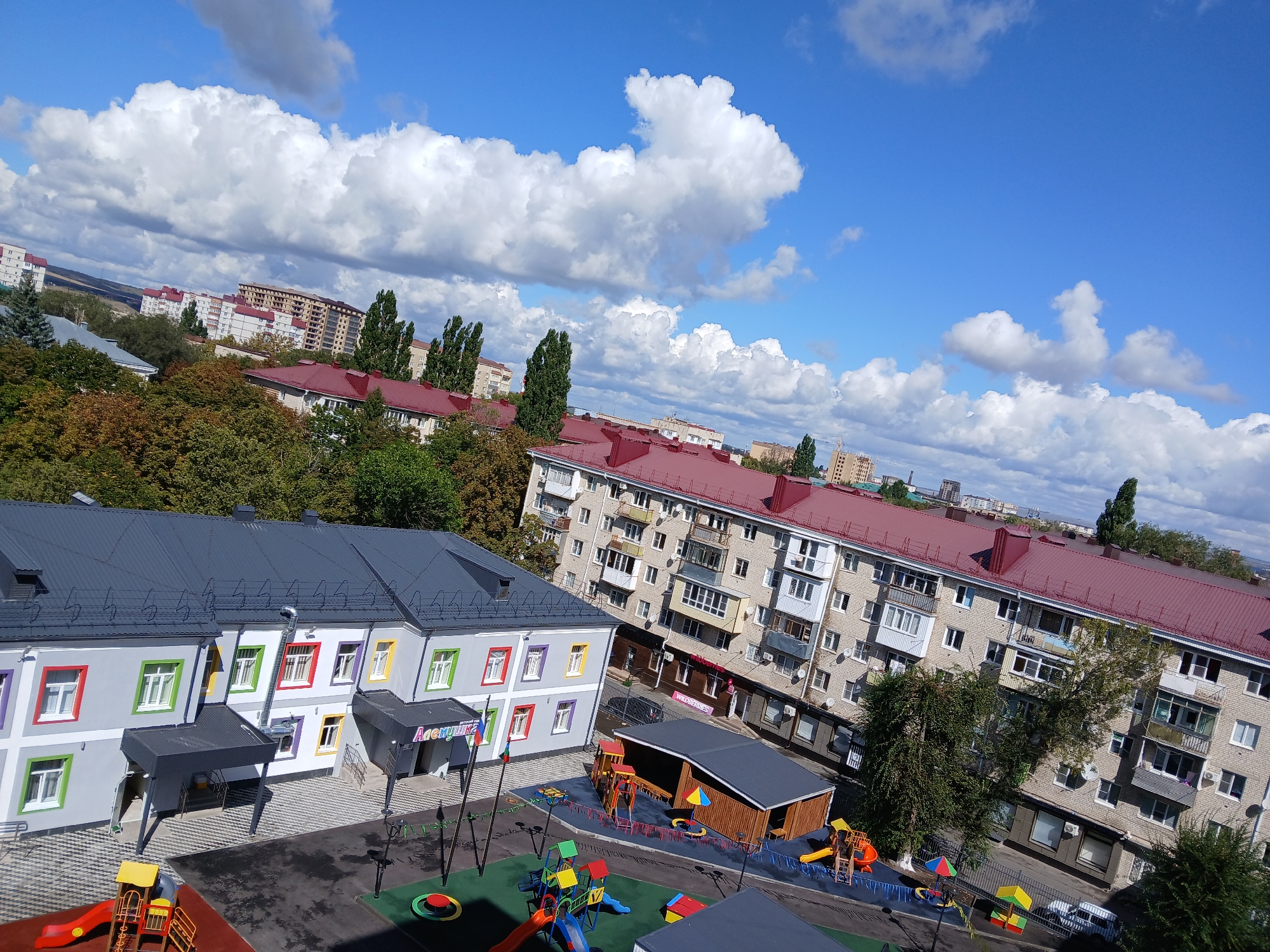 The aerial photograph captures a sunny day in Cherkessk, Russia, with a bright blue sky dotted with fluffy white clouds. The scene is dominated by urban architecture and a vibrant children's playground. In the foreground, a modern, two-story building with a gray roof and a facade painted in light gray with colorful window frames (red, green, yellow, purple, and white) stands prominently. Two dark gray awnings extend from the building's entrances. To the right of this building, a multi-story apartment complex with a red-tiled roof, light brick facade, and numerous balconies is visible. In front of the apartment complex, a large, well-equipped children's playground features slides, swings, climbing structures, and colorful play mats. A smaller wooden structure with a gray roof is also part of the playground area. Beyond these buildings, more apartment buildings and trees are scattered across the landscape, with a few taller buildings and a crane visible in the distance, indicating ongoing construction. The overall impression is a well-maintained urban environment with a focus on recreational facilities for children.