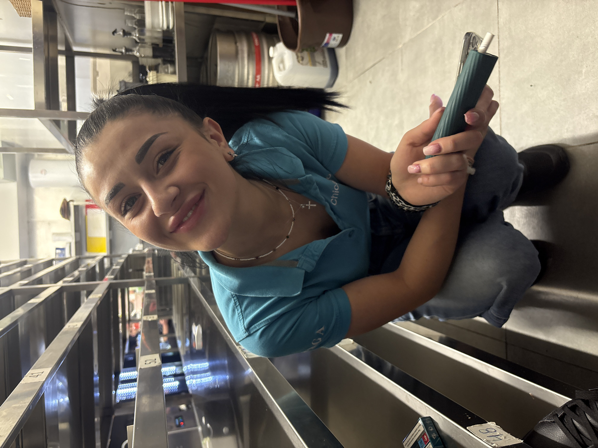 A young woman with long dark hair, wearing a blue polo shirt, is sitting and smiling at the camera. She is holding a green electronic cigarette in her hands. Her nails are painted with a pink ombre effect. She is wearing jeans, a beaded necklace with a cross pendant, and a black and white bracelet. She is sitting amidst metal shelves or drawers, possibly in a storage room or work area. In the background, there are kegs and containers on shelves. There is a partial view of what appears to be a shelving system with numbered slots. The location and city are unknown.