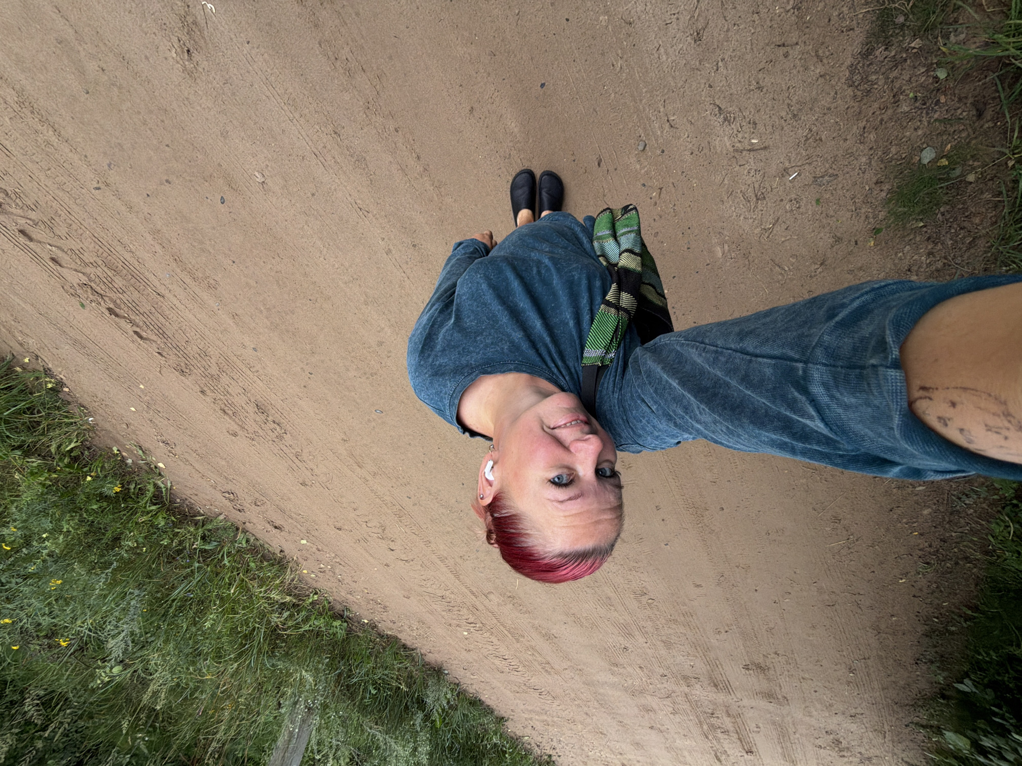 A woman with short, bright pink hair is taking a selfie while standing on a dirt path. She is wearing a teal long-sleeved shirt and black slip-on shoes. She has a patterned green and black bag slung across her body. Her left arm is extended towards the camera, holding the phone. She is smiling and looking directly at the camera. The path appears to be in a rural or park-like setting, with tall green grass and some yellow wildflowers on the left side of the path. There are tire tracks visible on the dirt path. The background is out of focus but suggests a natural environment. The image was taken in Chaikovsky, Russia.