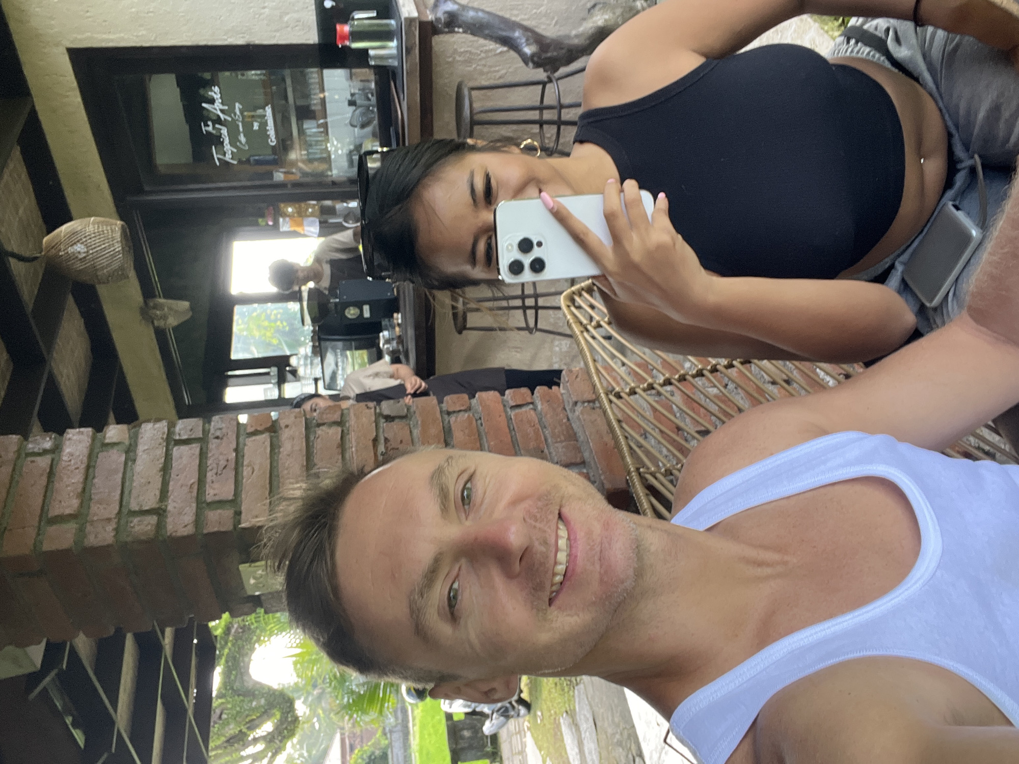 In Ubud District, Indonesia, two individuals, a man and a woman, are seated at a rustic outdoor cafe. The man, in a white tank top, smiles directly at the camera. The woman, wearing a black tank top, partially obscures her face with a white smartphone as she takes a selfie. Behind them, a barista works at a counter adorned with a sign that reads "The Tropical Apds Coffee and Eating by Goldmine." The cafe has a brick pillar, a woven light fixture, and visible greenery outside.