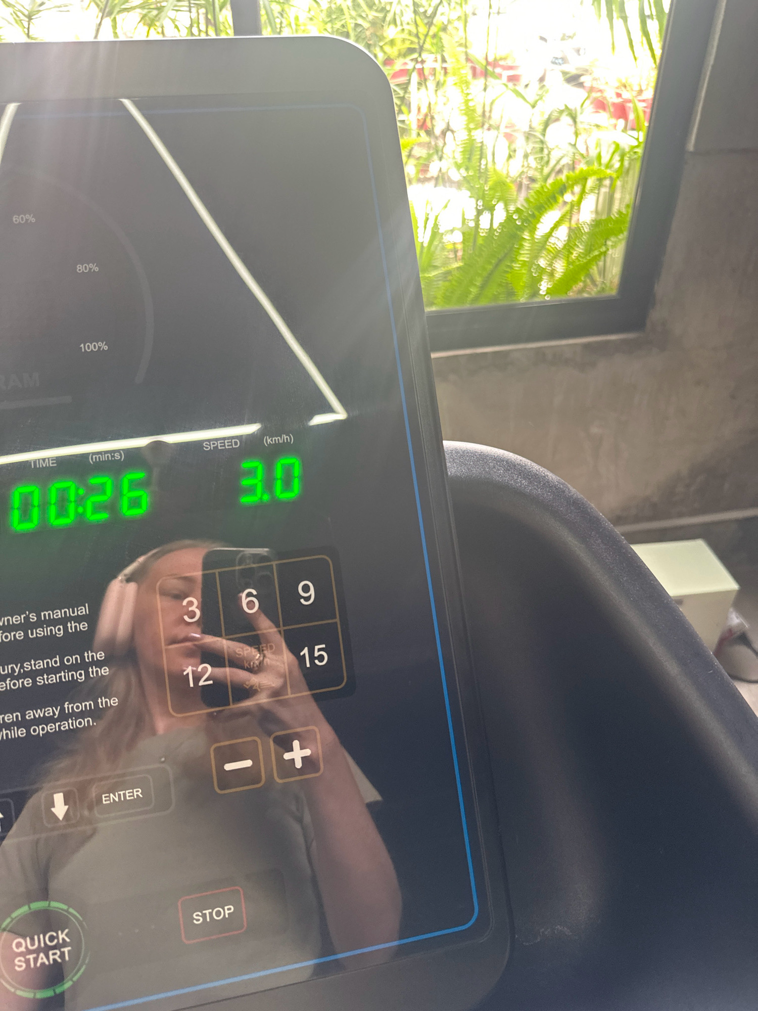 The image shows a close-up of a treadmill console in an indoor setting. The console displays the time as "00:26" and the speed as "3.0 km/h". Below the display, there is a numerical keypad with numbers 3, 6, 9, 12, and 15. There are also buttons for "-", "+", "ENTER", and a down arrow. A person's reflection is visible in the console, showing them wearing headphones and holding a phone. The background shows a window with lush green plants and sunlight filtering through. The scene appears to be a gym or fitness area. The location is not specified in the image.