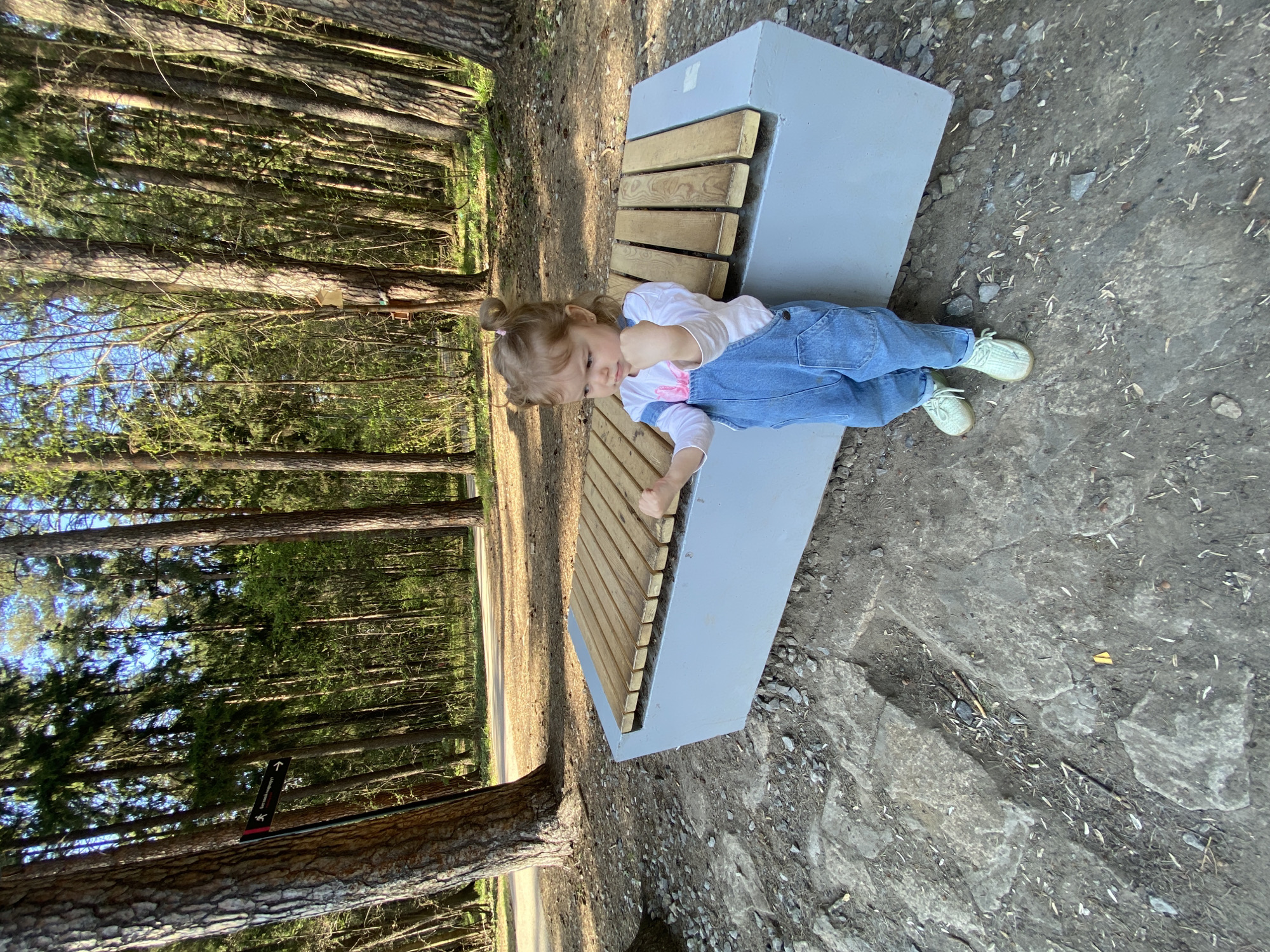 A young child is lying on a modern bench in a park setting. The bench is constructed of a light gray, possibly concrete, base with wooden slats forming the seating surface. The child is wearing blue denim overalls over a white long-sleeved shirt and light-colored shoes. The child's body is positioned diagonally across the bench, with their head near the top and feet near the bottom of the image.
The location appears to be a park on the edge of a forest. Tall, thin trees are visible behind the bench, stretching high into the sky. The ground around the bench is a mix of dirt and small rocks. Based on the park's style and bench design, the location is possibly in the city of Perm, Russia.