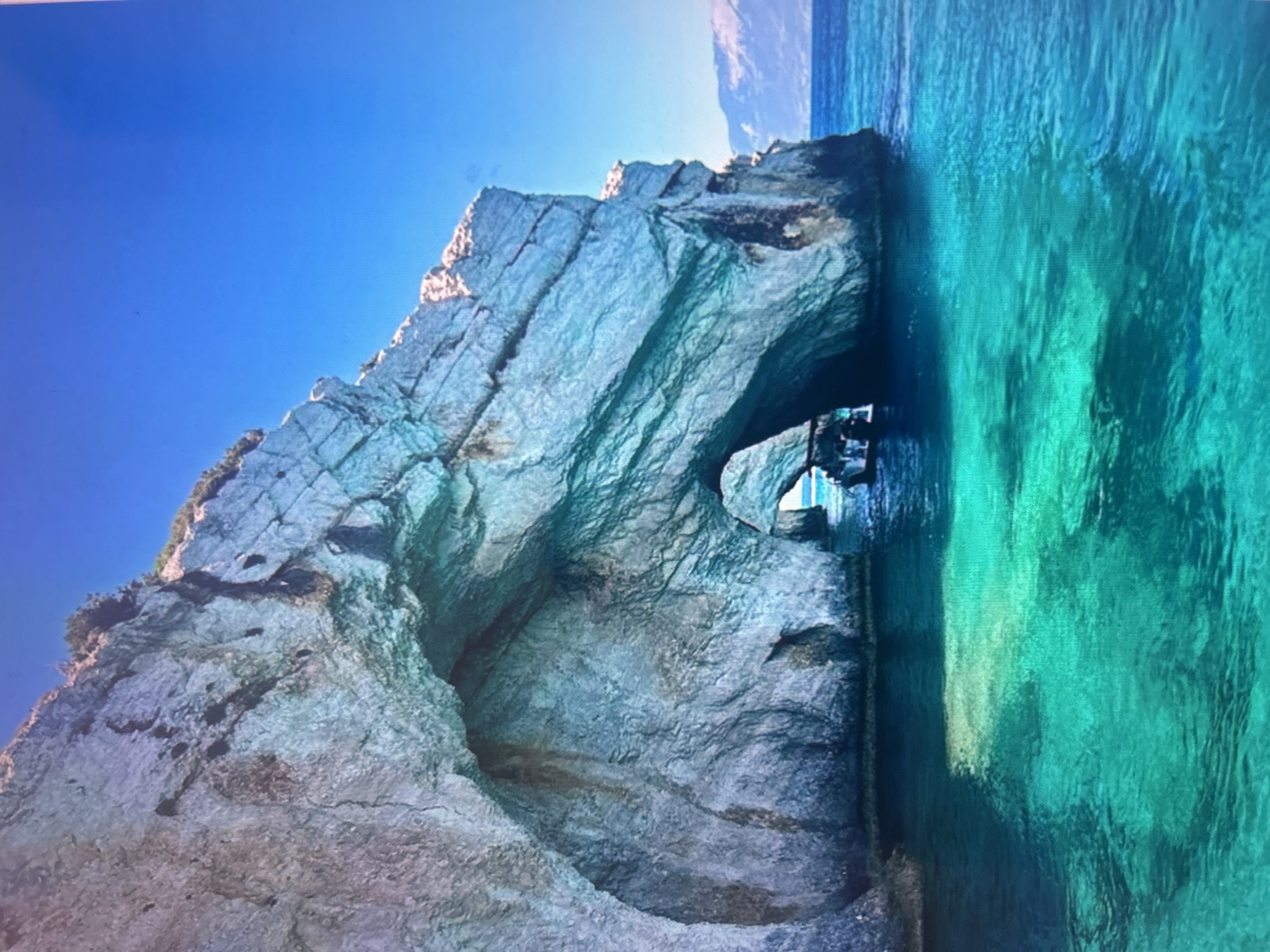 The image, presented rotated 90 degrees clockwise from its natural orientation, depicts a stunning coastal scene featuring a large rock formation and clear blue-green waters under a bright sky. If viewed correctly, the vast, cloudless blue sky would be at the top, and the vibrant water at the bottom, with a massive cliff face dominating the left side of the frame.
The central subject is a towering, weathered rock structure, likely limestone, characterized by its rugged texture, deep crevices, and natural fractures. Its surfaces display a range of light grey, white, and pale blue-green hues, intensely illuminated by bright sunlight on its upper and outer portions. At the base of this formation, a prominent natural archway or sea cave has been carved out by the sea. The entrance of this cave is wide and inviting, leading into shadowed depths where reflections of light play on the water. Within the cave's opening, dark shapes suggest the presence of a small boat, possibly with individuals, hinting at exploration or leisure activities.
The water is remarkably clear and calm, exhibiting brilliant shades of turquoise and emerald green. These colors shift in intensity and tone depending on depth and light penetration, creating a captivating visual texture with subtle ripples and refracted light. The clarity of the water allows glimpses of the submerged rock formations. The bright, direct sunlight on the scene indicates a midday or early afternoon setting with perfect, cloudless weather. The overall impression is one of pristine natural beauty and serenity in a picturesque coastal location. No visible text is present.