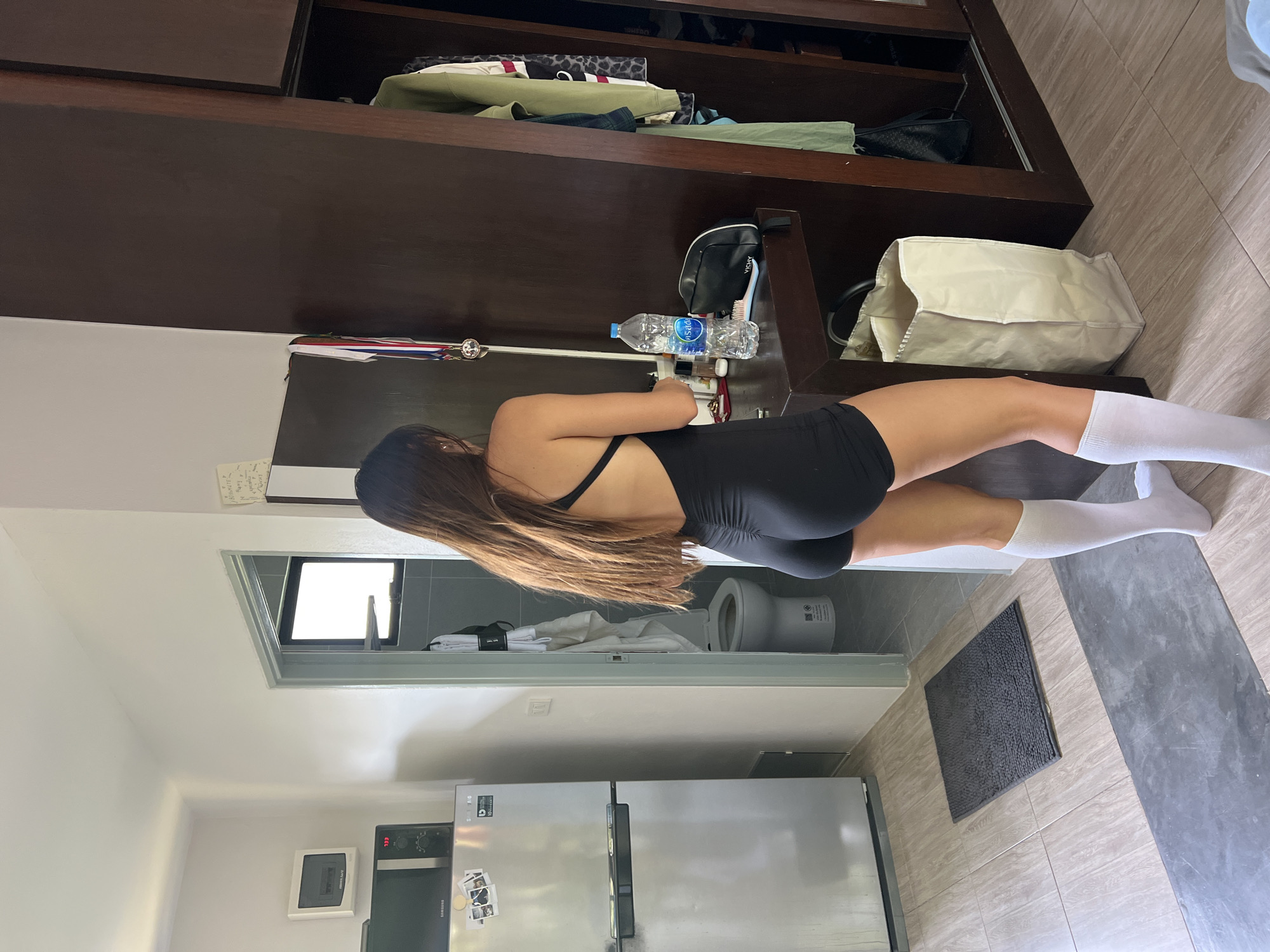 A woman with long brown hair is standing in a room in บ้านบางรักษ์, Thailand. She is wearing a black one-piece outfit and white socks. She appears to be reaching into a dark wood cabinet, where there is a water bottle, toiletry bag, and other items. Behind her is a bathroom with a toilet visible through the doorway. Beside the bathroom is a kitchen with a stainless steel refrigerator and microwave. There is a small mat on the floor.