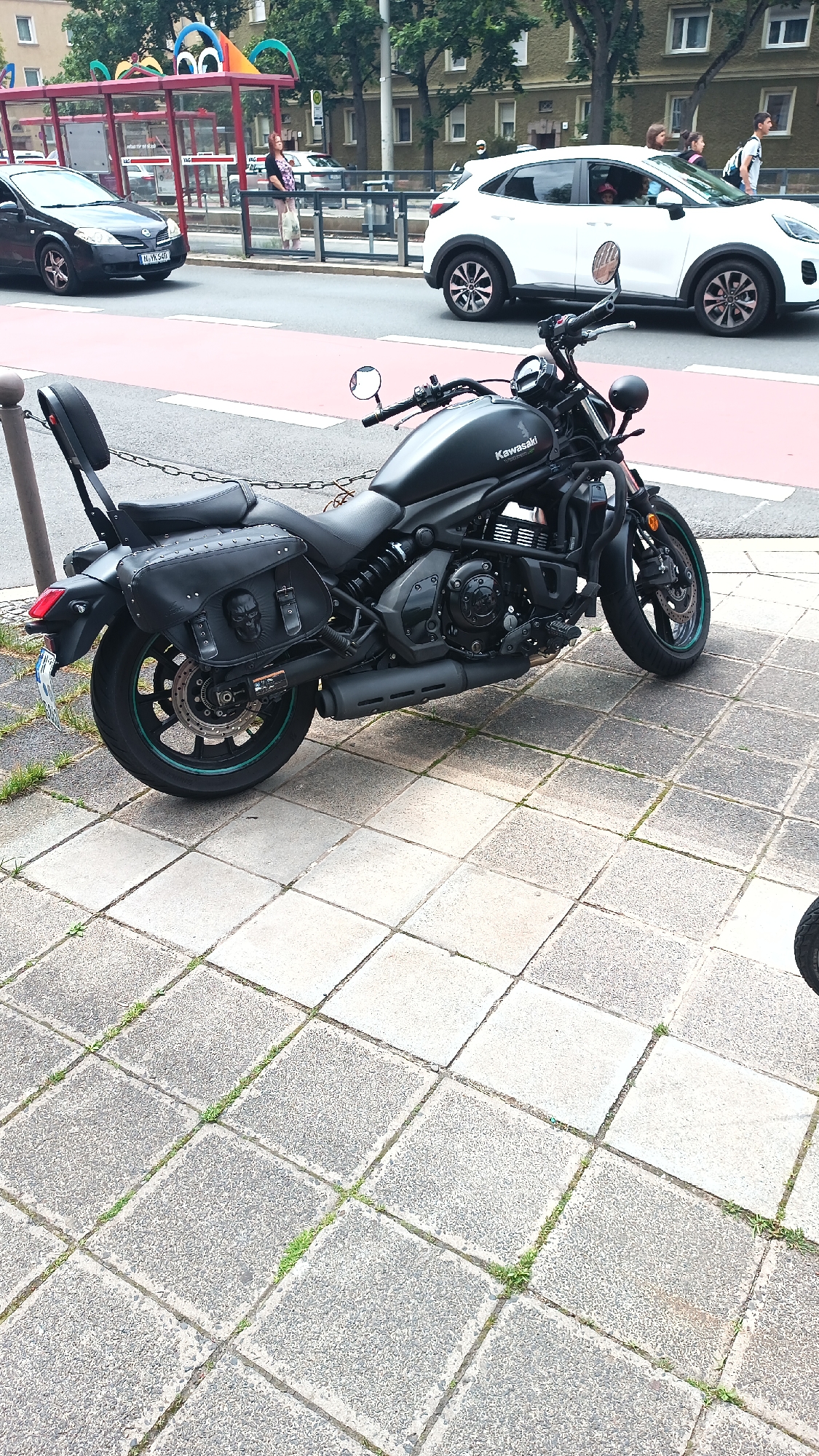 A black Kawasaki motorcycle is parked on a paved sidewalk in Nuremberg, Germany. The motorcycle has a black leather saddlebag with a skull design on it. In the background, a black car and a white SUV are driving on the street, and a bus stop with people waiting can be seen. The weather appears to be sunny with trees and buildings in the background.
