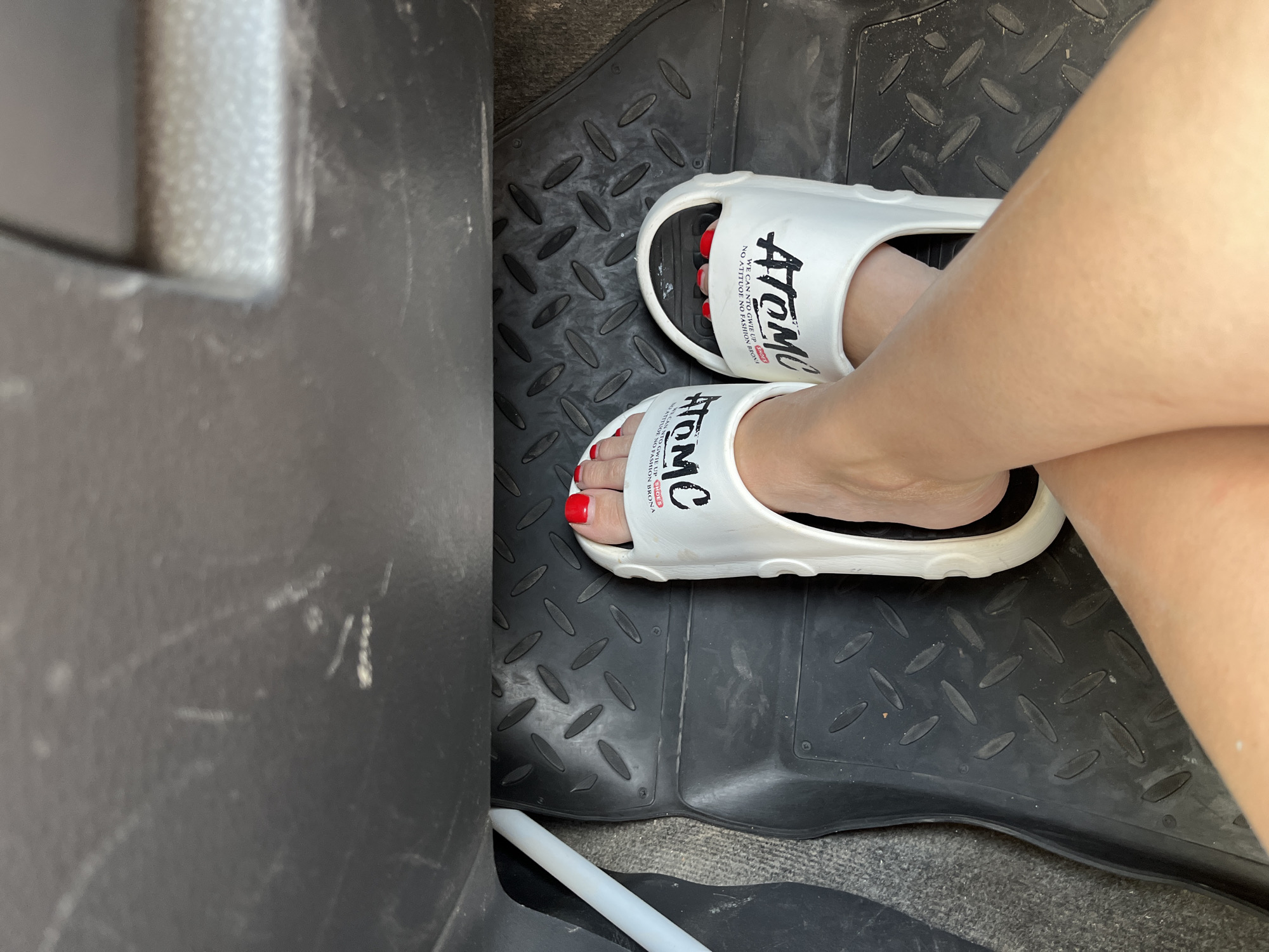 A pair of white flip-flops with black lettering are on a black rubber car mat. The lettering on the left flip-flop appears to read "ATOM" and "SHOES." The lettering on the right flip-flop is partially obscured, but also seems to read "ATOM" and "SHOES." The toes of a person wearing red nail polish are visible in the flip-flops. The feet are positioned in a car, with the car's interior visible. The location is stated to be in the city of Пристанное, Russia.