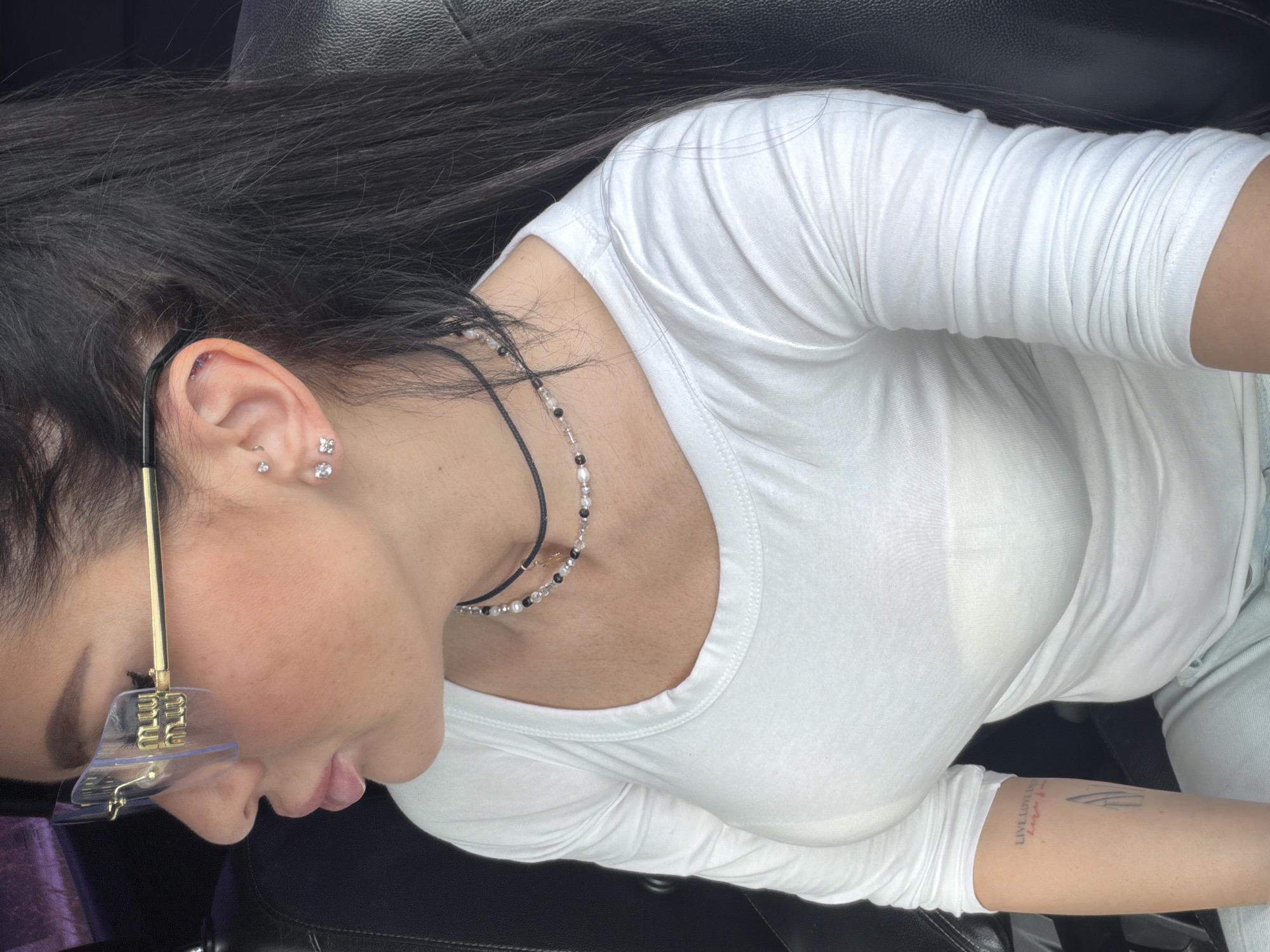 The image shows a person with dark hair wearing a white t-shirt. They are wearing unique sunglasses with a metallic frame and transparent attachments at the bottom. The person has several piercings in their ear and is wearing a two-strand necklace. A tattoo is visible on their inner forearm. The person is sitting in what appears to be a black leather car seat. There is no information to identify the location as Voronezh, Russia.