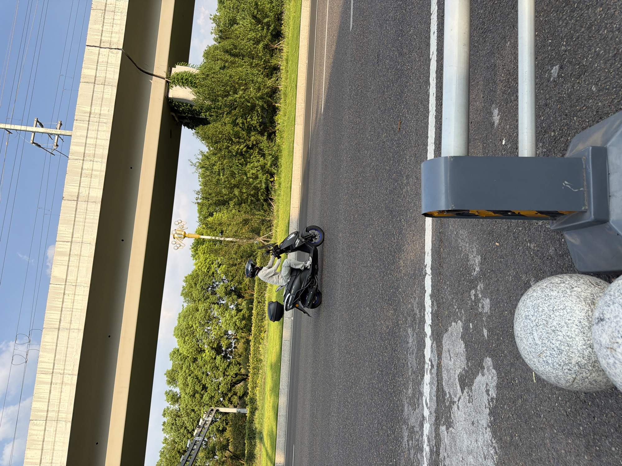 An individual wearing a light-colored long-sleeved shirt, grey pants, white sneakers, and a black helmet is riding a black scooter on a paved road. The scooter has a black storage box mounted on the rear. The road has white lane markings. In the background, there is a large concrete overpass with visible rebar and electrical wires running overhead. Lush green trees and vegetation line the side of the road beneath the overpass. The sky is blue with scattered clouds. In the foreground, there are two large, speckled grey granite spheres and a gray and yellow traffic barrier. This scene appears to be on a road in the Jiangjin District of Chongqing, China, likely near an elevated roadway.
