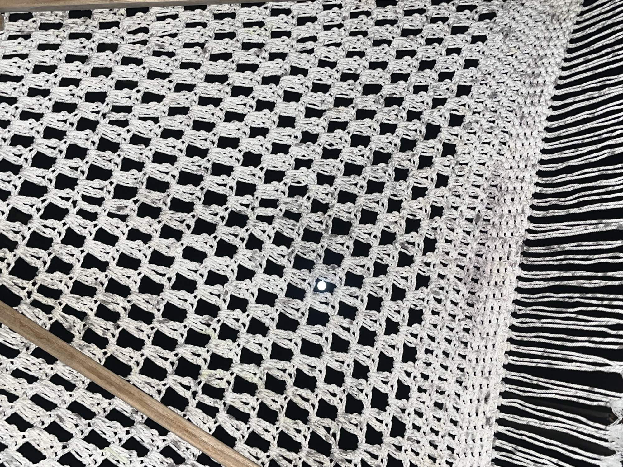 The image depicts a crocheted white hammock with fringed edges against a black background, possibly suspended or laid flat. A segment of a wooden beam is visible along the left edge. The scene is characterized by its high-contrast and focus on textile detail. There is no clear indication of the location being Kuta Selatan, Indonesia within the visual information provided.