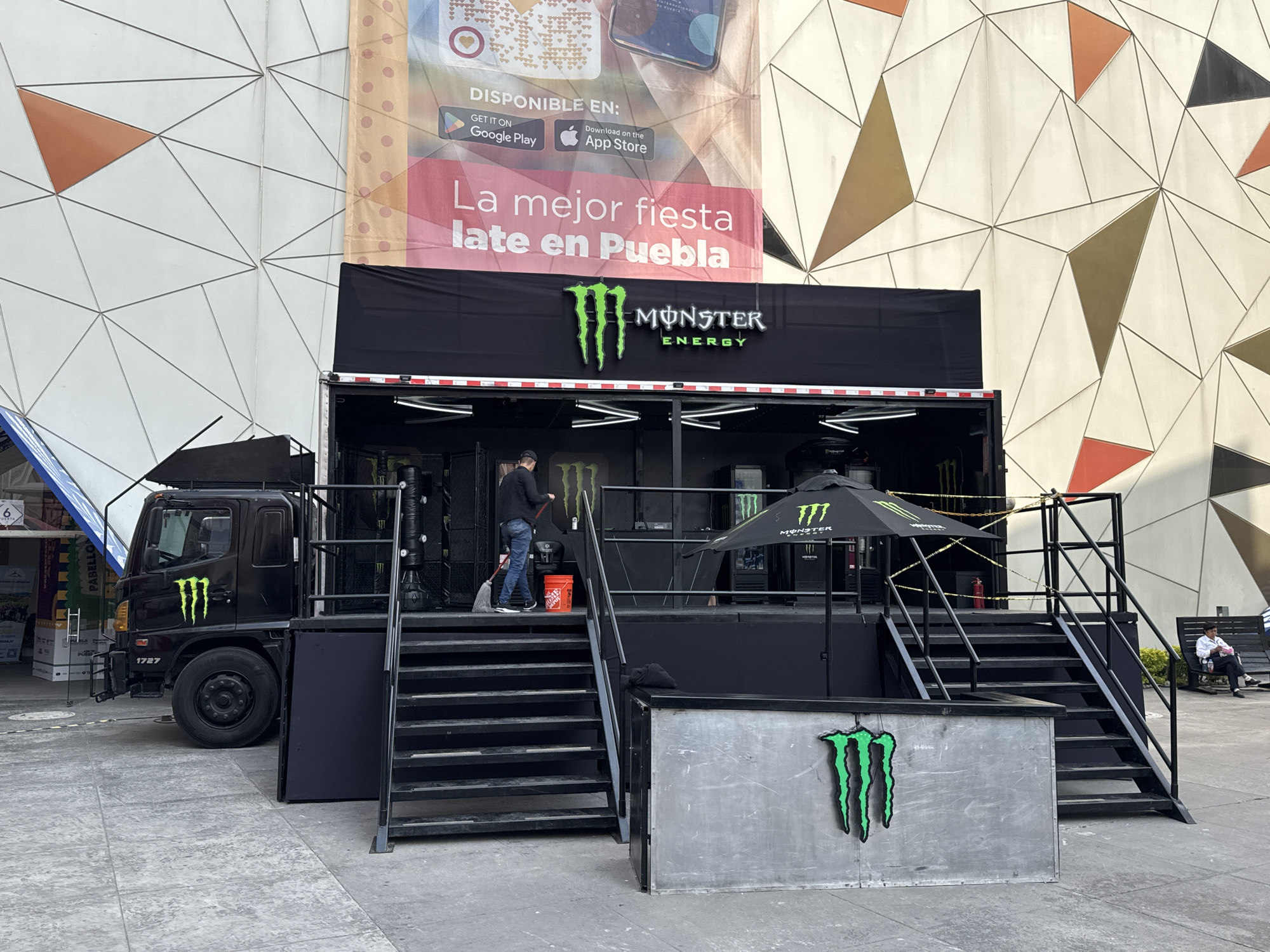 The image captures a Monster Energy marketing setup in what appears to be an outdoor public space in Puebla, Mexico. A black truck with the Monster Energy logo is attached to a small stage, also branded with Monster Energy. There are two sets of black metal stairs leading up to the stage on either side. A person is seen on the stairs near the truck, using a mop. An advertisement banner is visible behind the setup, advertising "La mejor fiesta late en Puebla" (The best late party in Puebla) and indicating app availability on Google Play and the App Store. The backdrop is a modern building facade with a geometric pattern of triangles in white, beige, and orange. The scene suggests a promotional event or activation for the Monster Energy drink in Puebla.