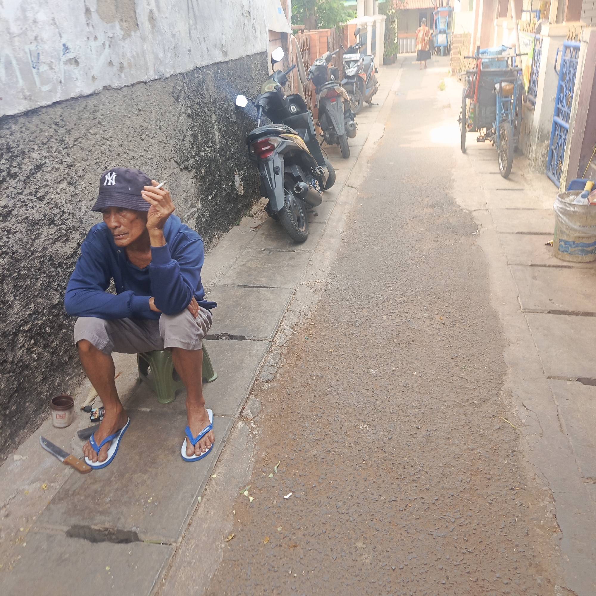 The image depicts a man sitting on a green plastic stool on a sidewalk next to a weathered, textured wall. He is wearing a blue hooded sweatshirt, light gray shorts, and blue and white flip-flops. On his head, he wears a dark blue bucket hat with a white "NY" logo. He is holding a cigarette in his right hand, near his mouth, and appears to be smoking. To his left, on the sidewalk, is a small brown container, a lighter, and a knife with a wooden handle.
The sidewalk is made of concrete slabs, some of which are cracked. The wall beside him is rough and has patches of discoloration. Behind the man, several motorcycles are parked along the sidewalk, indicating an urban setting. The street itself is paved with asphalt and appears to be a narrow alleyway, as suggested by the buildings and parked vehicles. In the background, a person in a patterned dress is walking away, and a three-wheeled cart is visible. The overall environment suggests a residential or commercial alleyway in an Indonesian city.