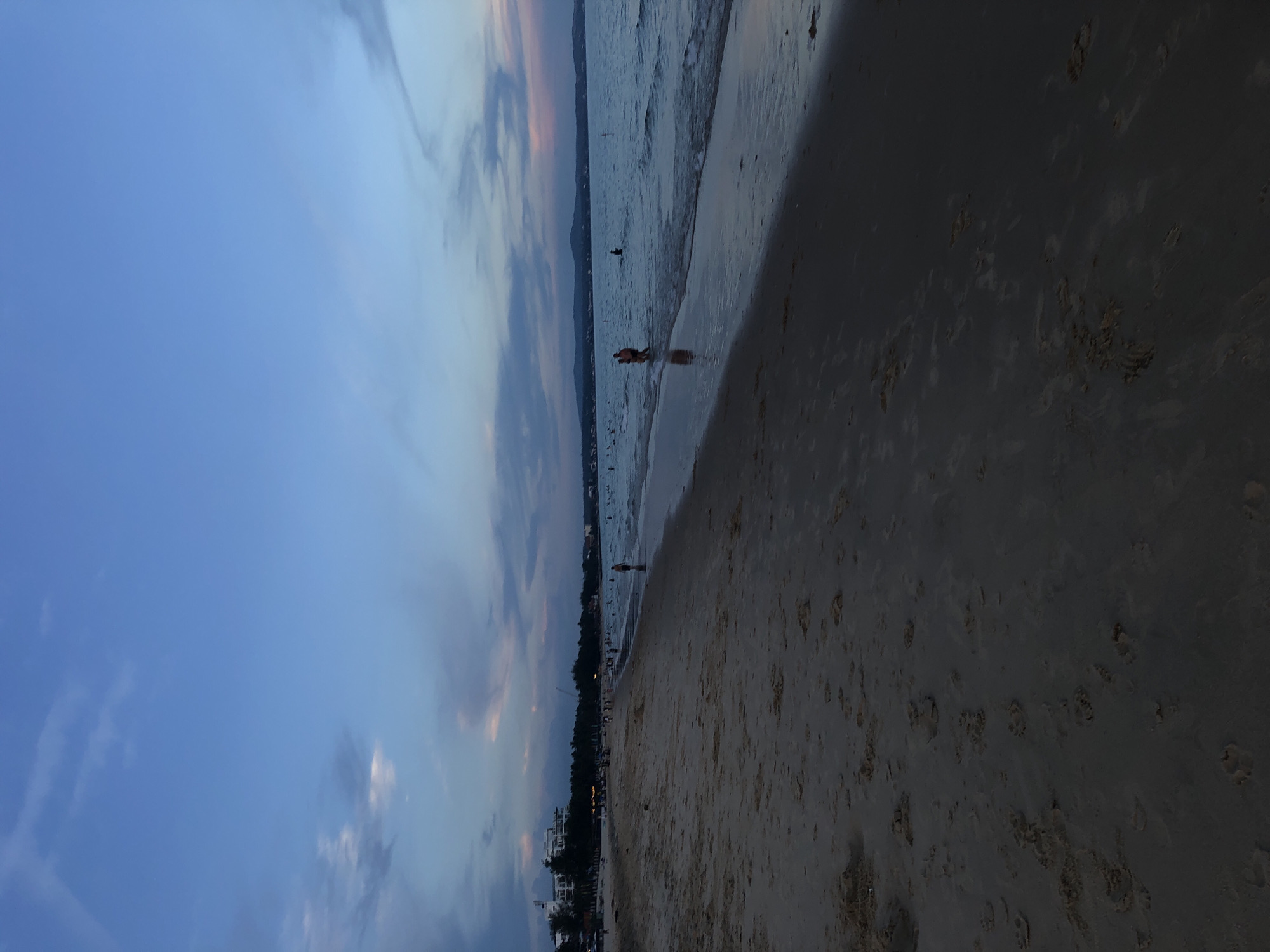 The image depicts a beach scene at dusk in Phu Thuy Ward, Vietnam. The sky is a gradient of deep blue transitioning to lighter blues and hints of pink and purple from the setting sun. Clouds are scattered across the sky. The beach is composed of light brown sand, with some footprints visible in the foreground. Gentle waves are washing ashore on the right side of the image. In the water, several people are present, some wading or swimming. Further in the distance, along the coastline, there are buildings that appear to be hotels or resorts, with lights beginning to illuminate. The overall atmosphere is tranquil, indicative of the end of a day at the beach.