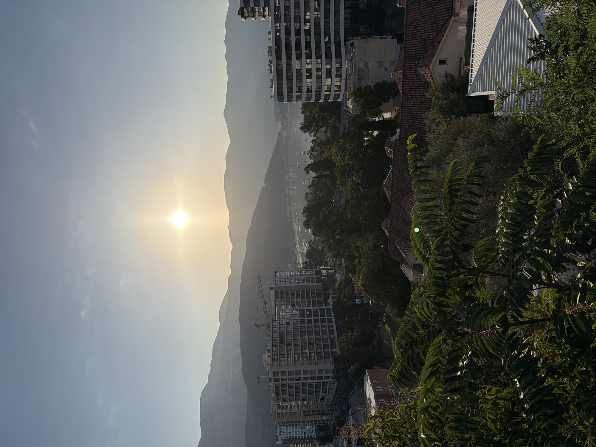The image depicts a scenic view of Budva, Montenegro, at sunset. The sun is low in the sky, casting a warm glow over the landscape. In the foreground, lush green foliage frames the scene, with hints of red-tiled roofs of houses visible.
Dominating the midground are several modern apartment buildings, some of which appear to be under construction, indicated by the presence of cranes. These buildings are characterized by their contemporary architecture, featuring numerous windows that reflect the sunlight.
Beyond the buildings, a body of water, likely the Adriatic Sea, is visible, dotted with several boats and yachts. The water leads to a picturesque bay.
In the background, a range of hazy mountains creates a dramatic silhouette against the sky. The sky itself is a soft blue, transitioning to a lighter hue near the horizon where the sun is setting. The overall impression is one of a tranquil coastal city bathed in the golden light of the setting sun, with a blend of modern development and natural beauty.
