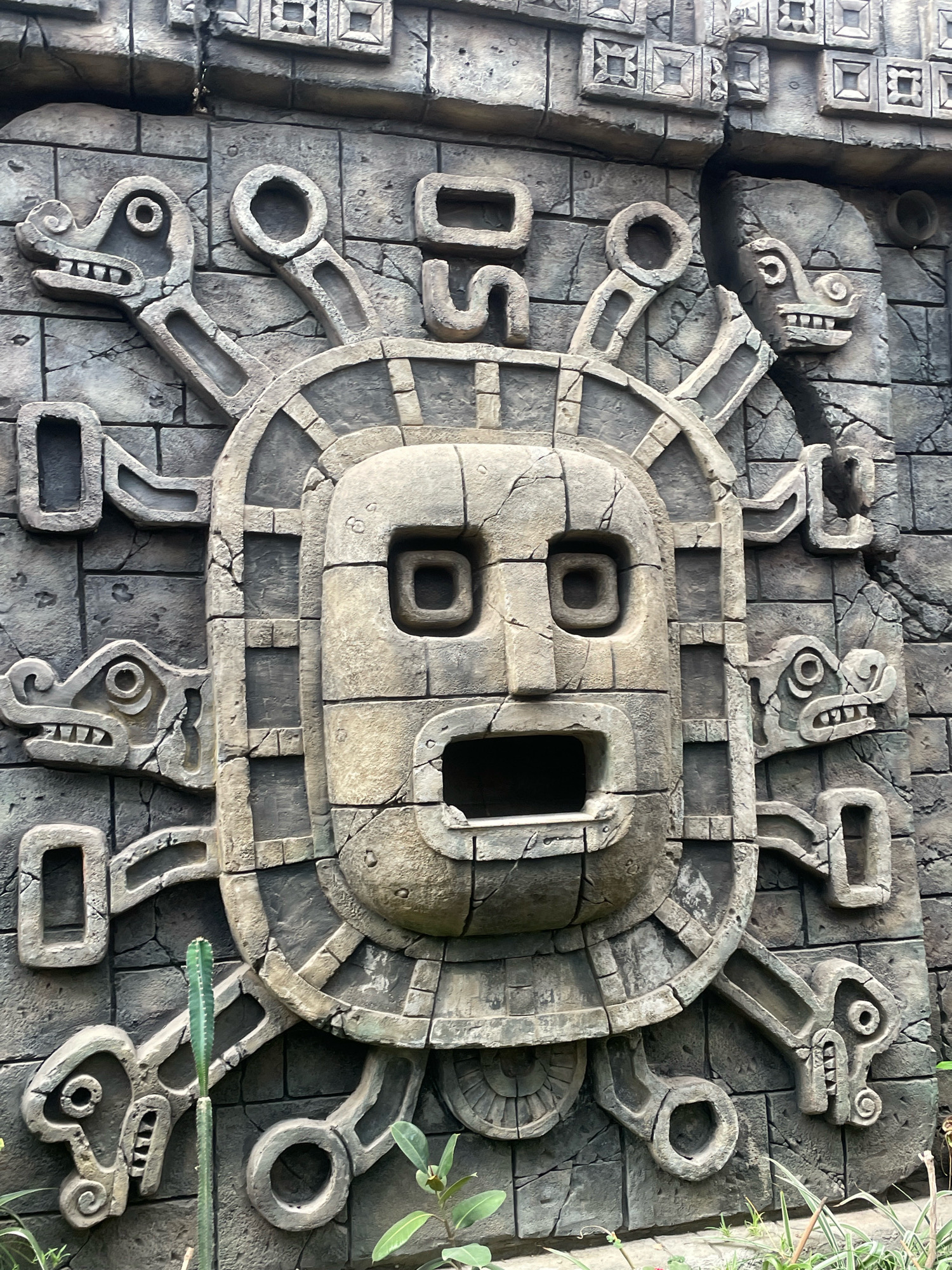 This is a detailed, close-up photograph of a large, weathered stone carving, likely a relief or part of an ancient facade. The carving depicts a stylized, monstrous face at its center, framed by a circular border. This border is adorned with multiple serpent-like heads with open mouths and sharp teeth, radiating outwards. The central face has large, square eye sockets, a gaping rectangular mouth, and a blocky nose. The entire structure is set against a background of intricately cut stone blocks, many of which are cracked and show signs of age and wear. The color palette is dominated by shades of gray and brown, reflecting the stone material. In the lower portion of the image, a thin, green cactus stands vertically in front of the carving, and some foliage is visible at the very bottom, suggesting an outdoor environment. The carving appears to be a significant piece of stonework, possibly representing a deity or a mythological creature. The image lacks any identifiable human presence or activity. There is no indication of this carving's location in the city of Krasnodar, Russia.