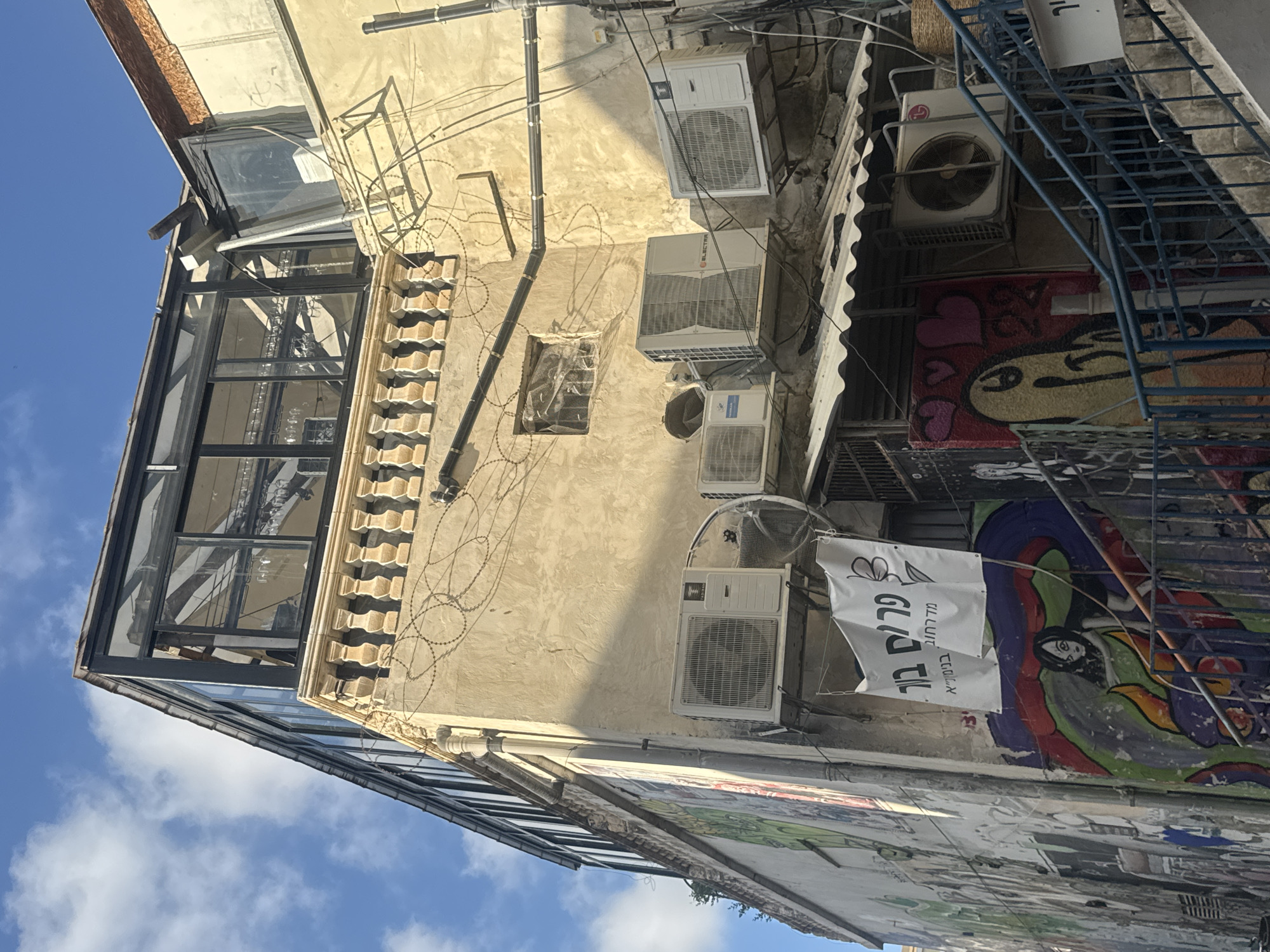 The image captures the exterior of a building in Tel Aviv, Israel, at what appears to be a narrow alleyway. The building features a tan-colored, textured facade with multiple air conditioning units mounted on the walls. A prominent element is a glass-enclosed structure on the upper level, with a decorative balustrade below it. Barbed wire is visible strung along the building's exterior, seemingly for security or to deter birds.
Graffiti art adorns the lower portion of the building, featuring colorful murals and stylized figures. A white banner with Hebrew writing and a butterfly graphic hangs prominently in the scene. A metal staircase with blue railings is visible on the right side, leading upwards. The sky above is blue with scattered white clouds. The overall impression is one of urban density and a mix of architectural styles, from the older building facade to the modern glass addition and the artistic graffiti.