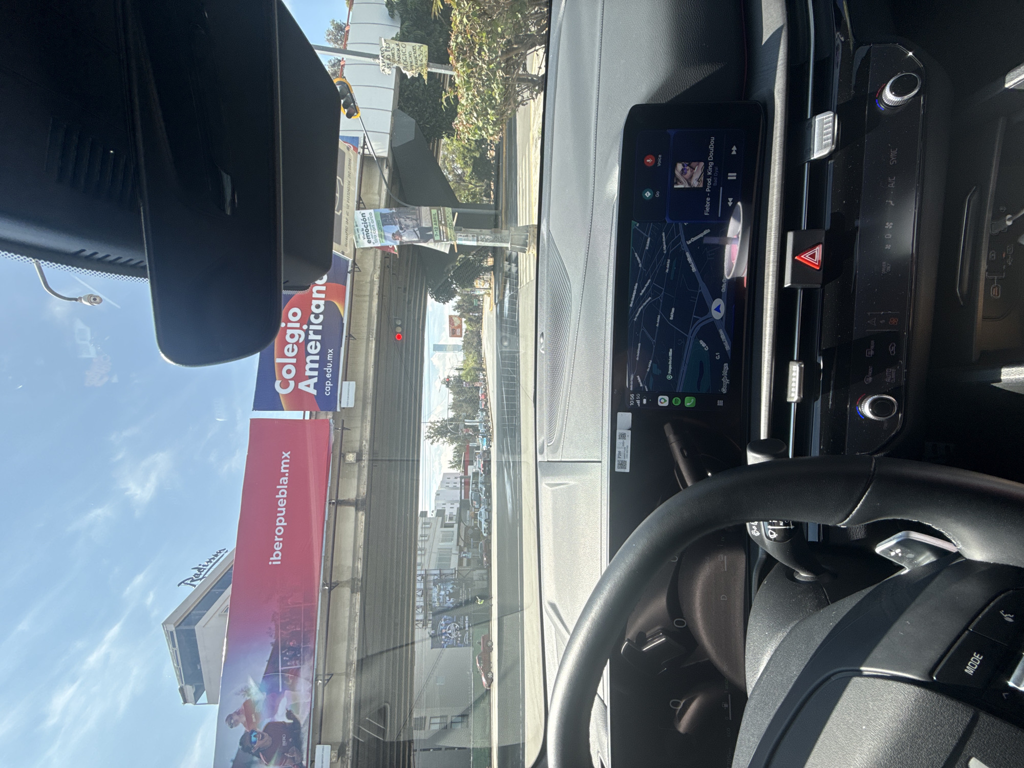 The view from inside a car on a road in Puebla, Mexico shows a cityscape dominated by a highway overpass and billboards. A large billboard advertises "iberopuebla.mx" and another promotes "Colegio Americano cop.edu.mx." A third advertisement says "Radisson." The car's dashboard displays a navigation app and music playing "Fiebre - Prod. King DouDou." The traffic light ahead is red. Road signs are visible in the background. A billboard with the words "RIMATO CARES PARQUE MOGICO SERVICIOS 163.24 CI" can be seen on the other side of the highway overpass.
