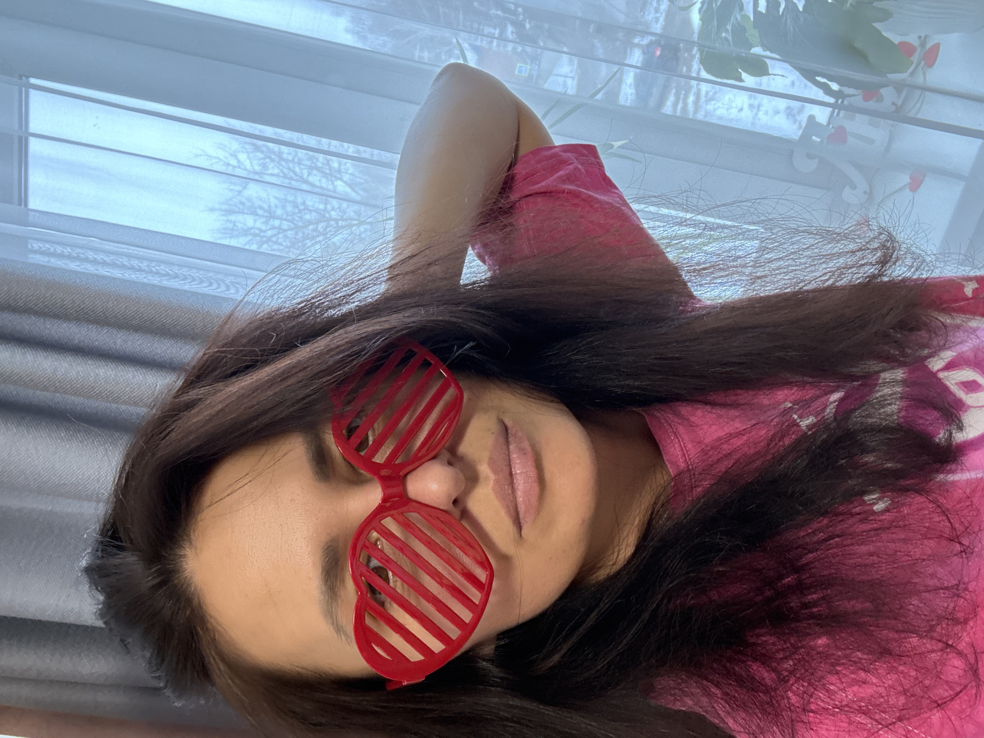 A woman with dark hair is wearing red heart-shaped sunglasses in a selfie. She is wearing a pink shirt and lying down with her arm above her head. The background shows a window with white blinds. The image appears to have been taken indoors during the daytime. The location of Chaikovsky, Russia is not explicitly confirmed.