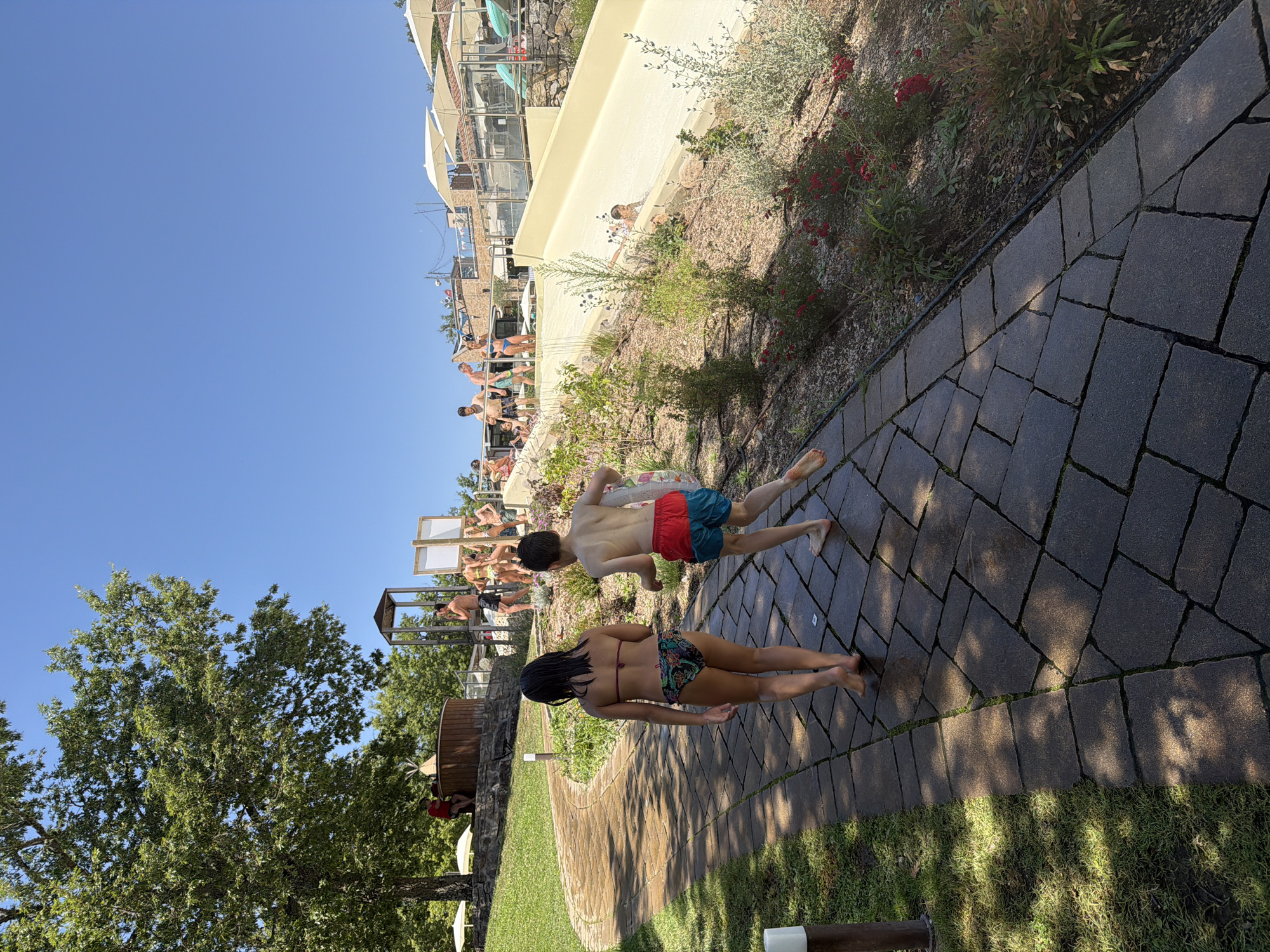 The image depicts a sunny day at a water park in Cavriglia, Italy. In the foreground, a young girl and boy walk away from the camera on a paved pathway. The girl wears a bikini with a patterned bottom and the boy wears red and blue swim trunks. To the right of the pathway, a water slide curves down a grassy slope, with a child in the process of sliding down. In the background, several adults and children are visible near the top of the water slide and around a wooden hot tub. The environment includes lush green trees, manicured lawns, and what appear to be buildings with terracotta roofs in the distance. The sky is a clear, bright blue.