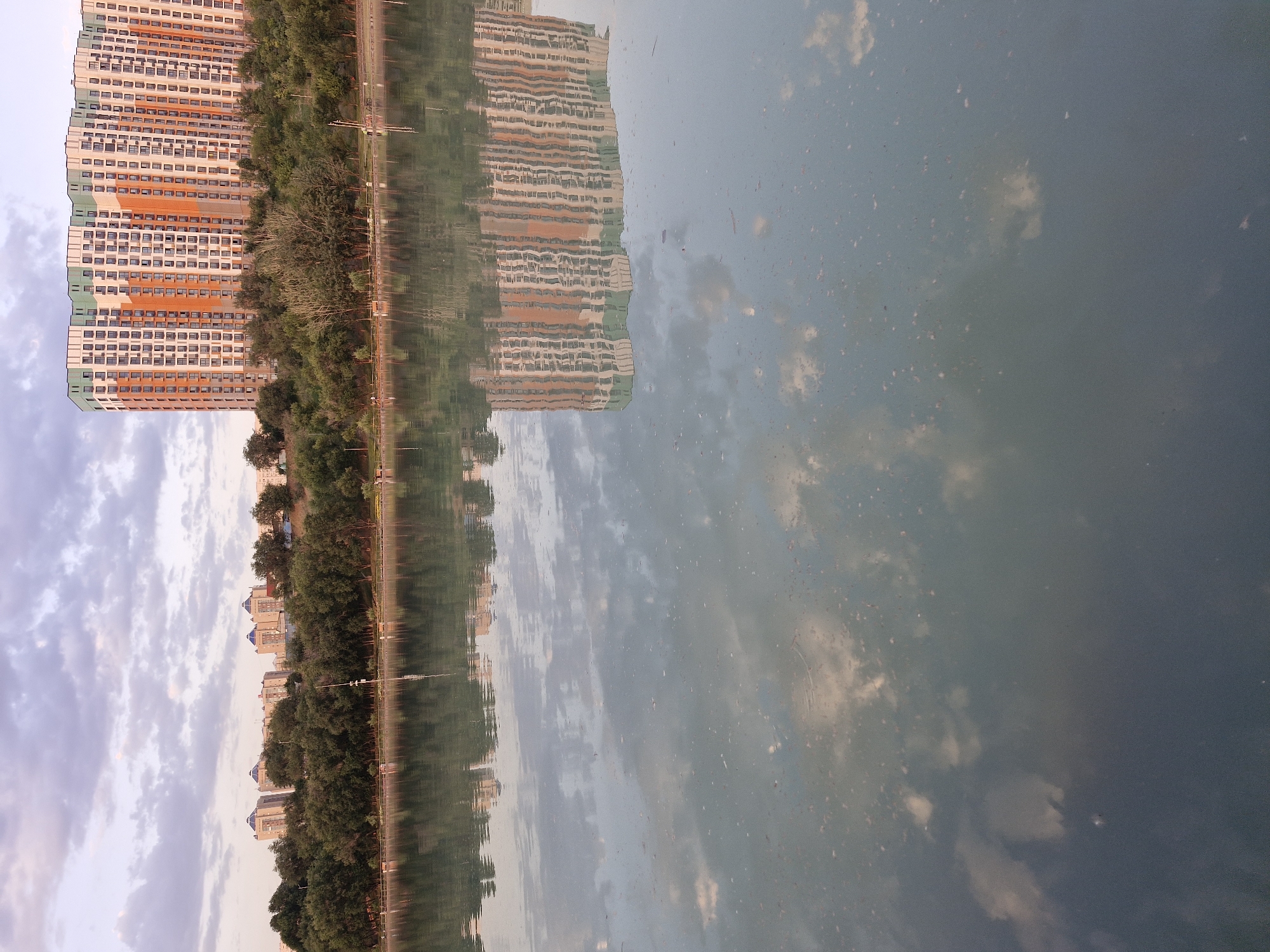 The image depicts a body of water with reflections of buildings and trees. In the background, a row of trees lines the shore, with several multi-story residential buildings visible beyond them. One prominent building, with a curved facade and alternating orange and white sections, is particularly noticeable. The water's surface is relatively calm, showing clear reflections of the sky, clouds, trees, and buildings. There are small particles scattered across the water's surface. The scene appears to be a park or recreational area within Almaty, Kazakhstan, likely during the late afternoon or early evening, given the soft lighting and the colors in the sky.