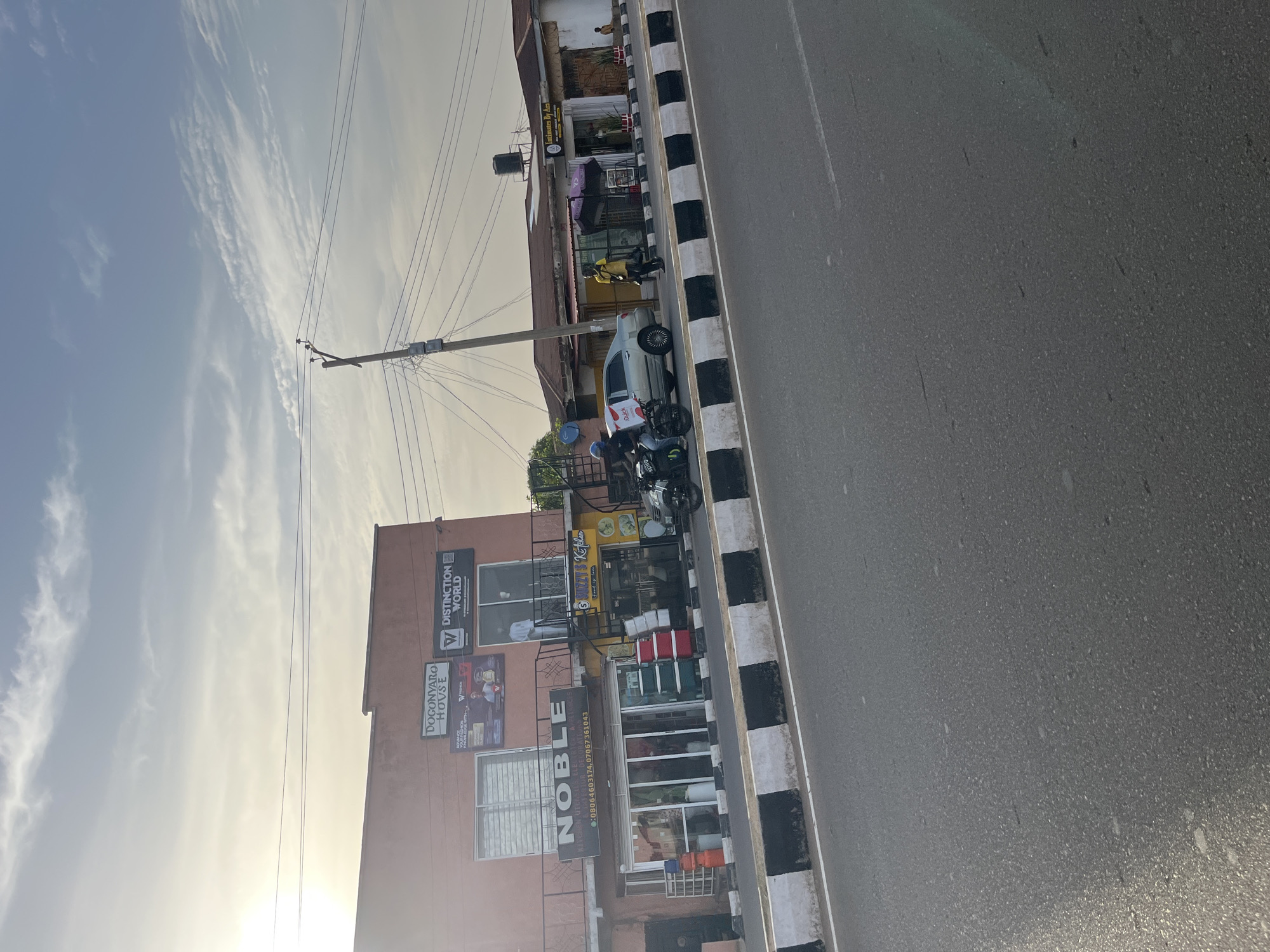 The image captures a street scene during what appears to be late afternoon or early morning, indicated by the soft, directional light and the low position of the sun creating a slight haze. The sky is light blue with wispy white clouds.
The main subjects are a road, a sidewalk/shoulder, and commercial buildings. The road, which is asphalt, runs horizontally on the right side of the frame, separated from the sidewalk by a black and white striped curb or median.
Along the left side of the road are several parked vehicles, including multiple motorcycles or scooters, one of which has a partially visible "Jumia" delivery box, and a white car.
Beyond the vehicles, a row of buildings lines the street. In the foreground, there's a reddish-brown multi-story building featuring several signs. Visible text on this building includes: "DISTINCTION WORLD" with a "W" logo; "DOGONYARO HOUSE" with "V VISIONS" and smaller text about "AUTHORS... KNOWLEDGE"; and prominently, "NOBLE" with phone numbers "08064603174, 07067361043". Smaller text below "NOBLE" suggests it offers "KITCHEN UTENSILS" and "HOME APPLIANCES". Above a yellow storefront next to this building, a sign reads "KITCHEN" or "SHIZZAY KITCHEN".
Further down the street, another building is visible, bearing a sign that reads "INTIMATES BY AXA". A large black water tank is perched on the roof of one of the buildings. A tall utility pole with numerous overhead power lines stands in the middle distance, extending into the sky.
A few individuals are visible in the scene: one person in a yellow top is walking near the curb, and another appears to be standing near a parked car. The activity is typical of a street, with parked vehicles and people moving about, suggesting a commercial area. The overall impression is one of a busy urban or semi-urban environment under fair weather conditions.
