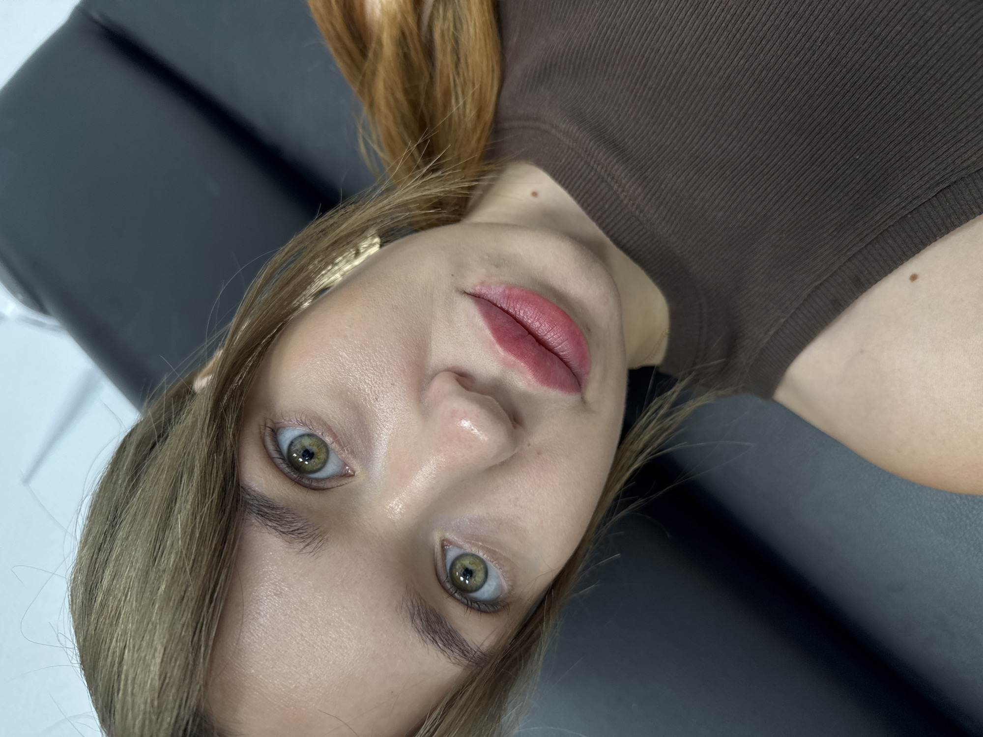 A close-up, high-angle, selfie-style shot focuses on the face of a young Caucasian female with light brown hair and green eyes. She is wearing a brown ribbed tank top. Her lips appear to be made up with a matte pink lipstick. The background is dark and out of focus, suggesting an indoor setting. The image does not provide any specific information about its location in the city of Chaikovsky, Russia, or any environmental context that would suggest a particular urban or natural setting. The lighting is direct and highlights the subject's skin, which appears to have a dewy finish. A small mole is visible on her collarbone.